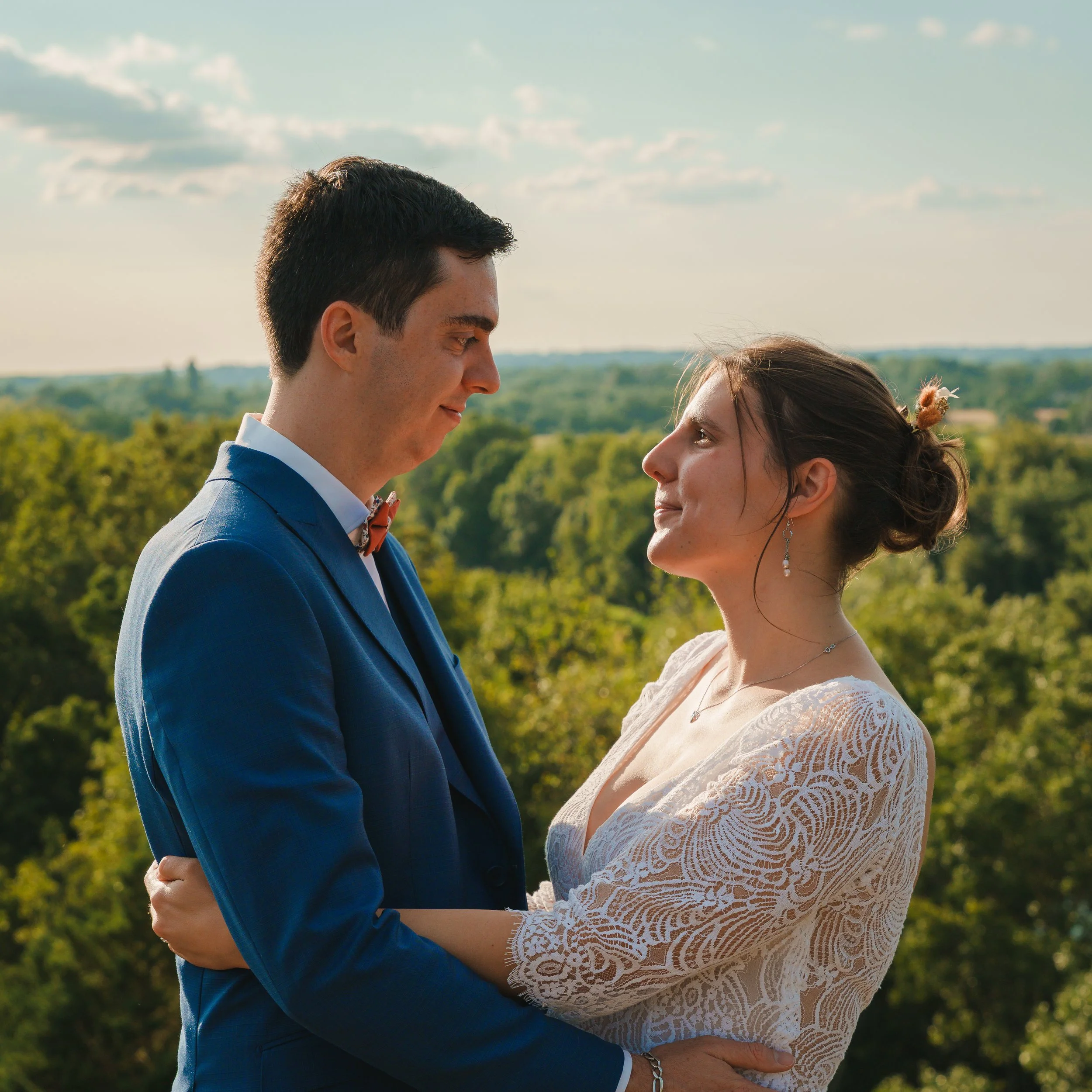 Un couple en tenue élégante se regarde tendrement dans un paysage verdoyant durant une journée ensoleillée.