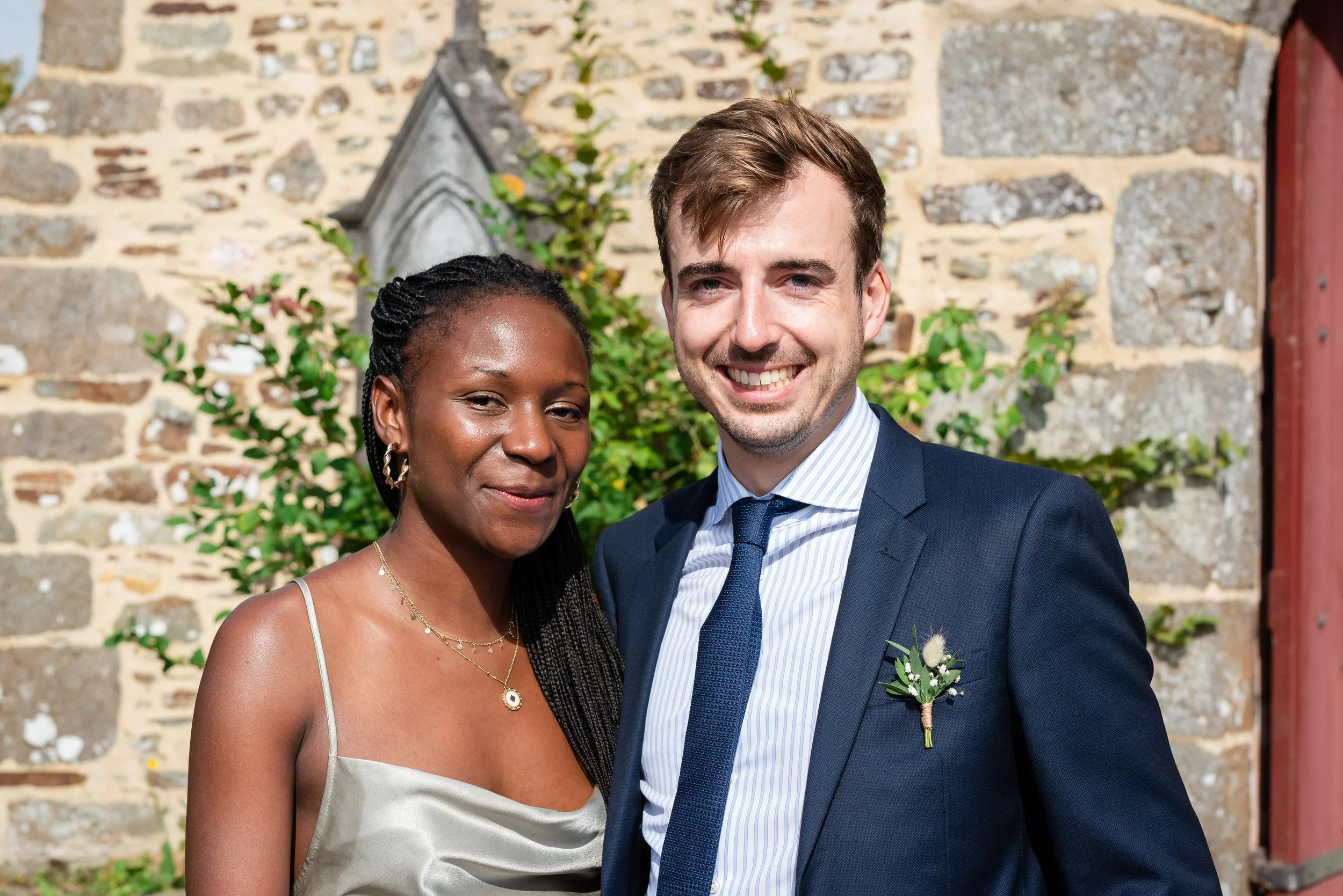 Deux personnes souriantes, un homme en costume et une femme en robe de soirée, posant devant un mur en pierre avec des plantes vertes, lors d'un événement ou d'une célébration.