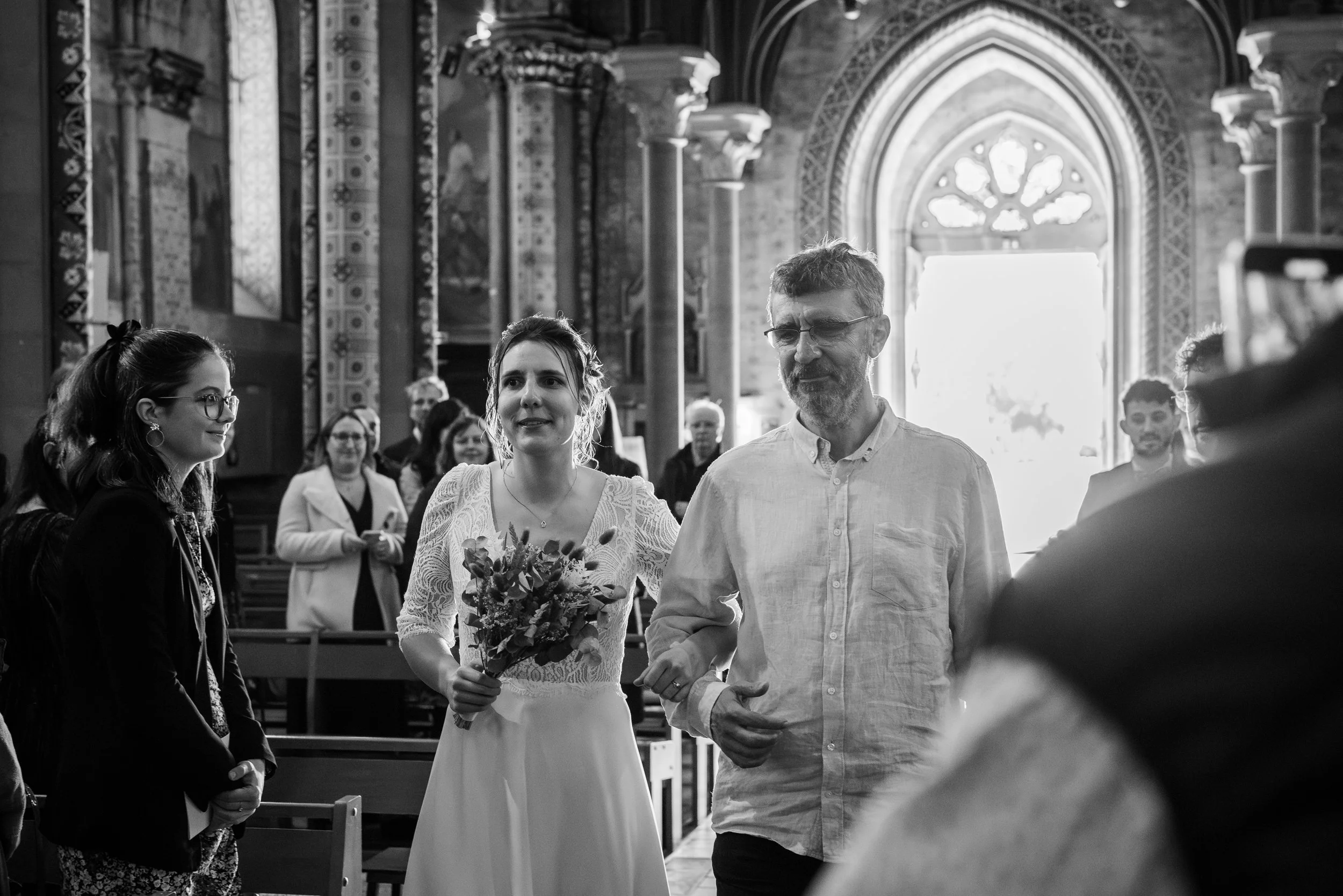 Une femme en robe blanche tient un bouquet de fleurs lors d'une cérémonie dans une église avec plusieurs personnes en arrière-plan.