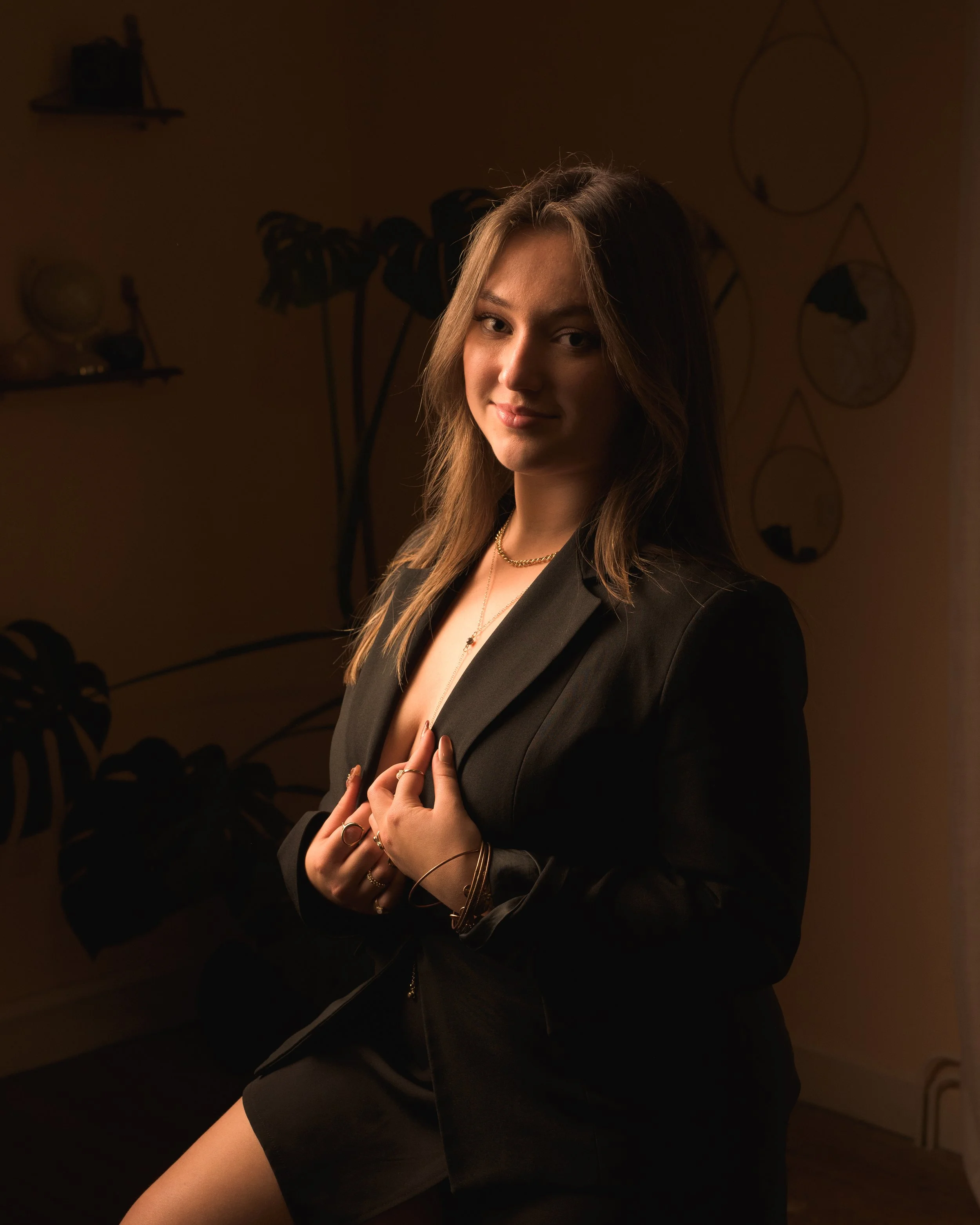 Une femme avec des cheveux longs et bruns, portant un blazer noir et des bijoux, se tenant dans un intérieur sombre, éclairée doucement.