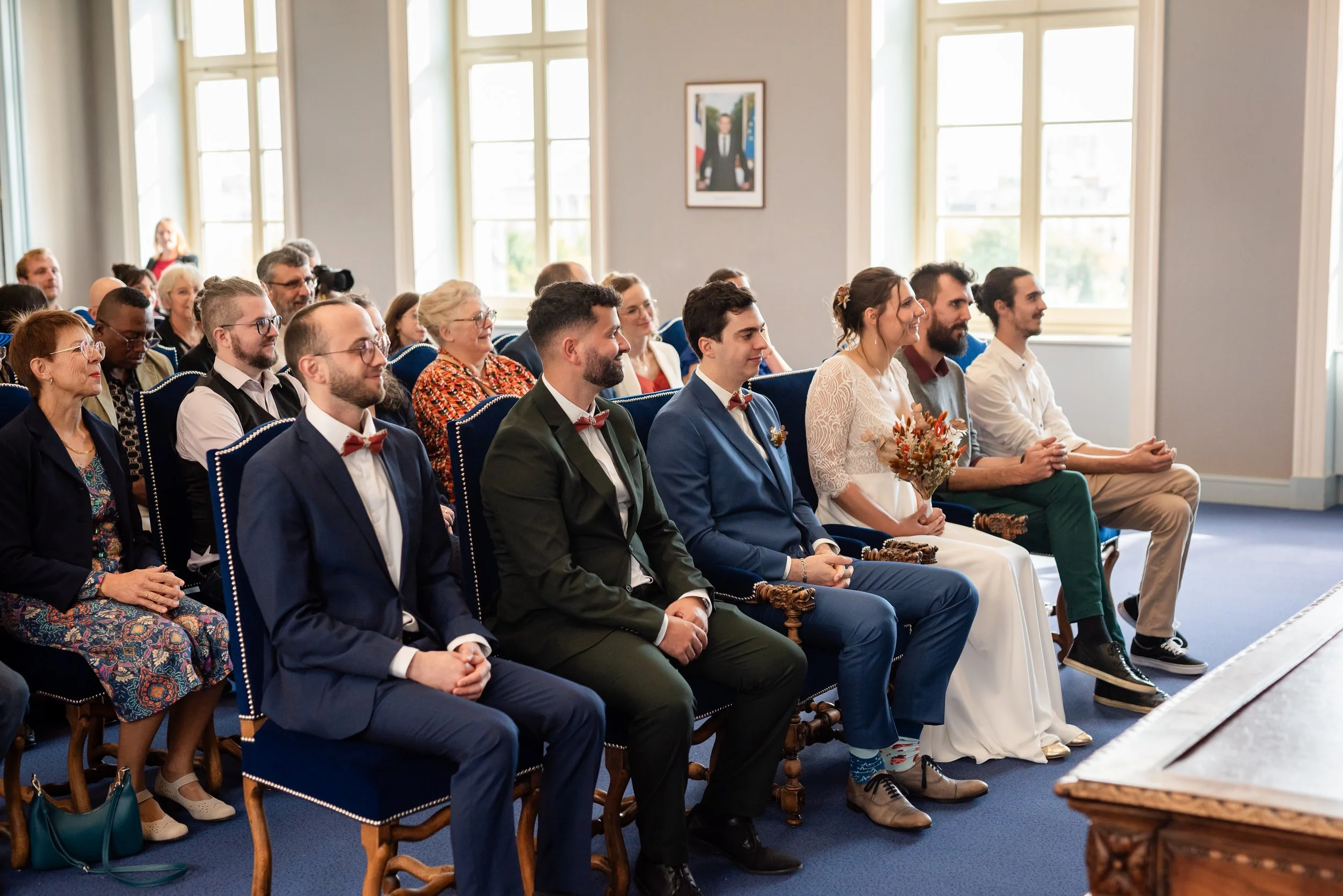 Un groupe de personnes assises dans une salle lors d'un événement formel, probablement un mariage ou une cérémonie officielle, avec des fenêtres grandes et lumineuses en arrière-plan.