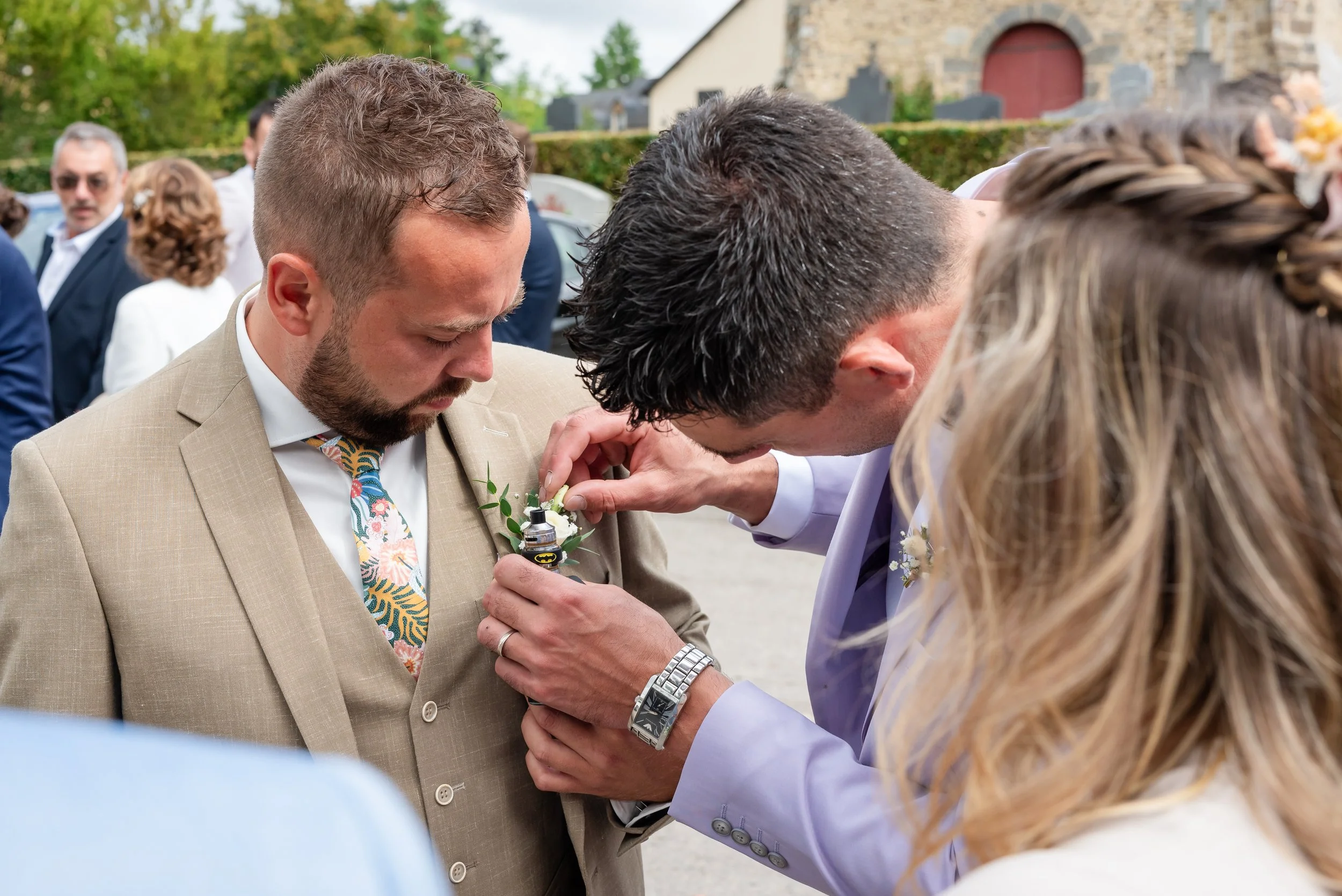 Un homme en costume beige avec une cravate colorée se voit remettre un objet par un autre homme lors d'une cérémonie en plein air, avec d'autres personnes en arrière-plan.