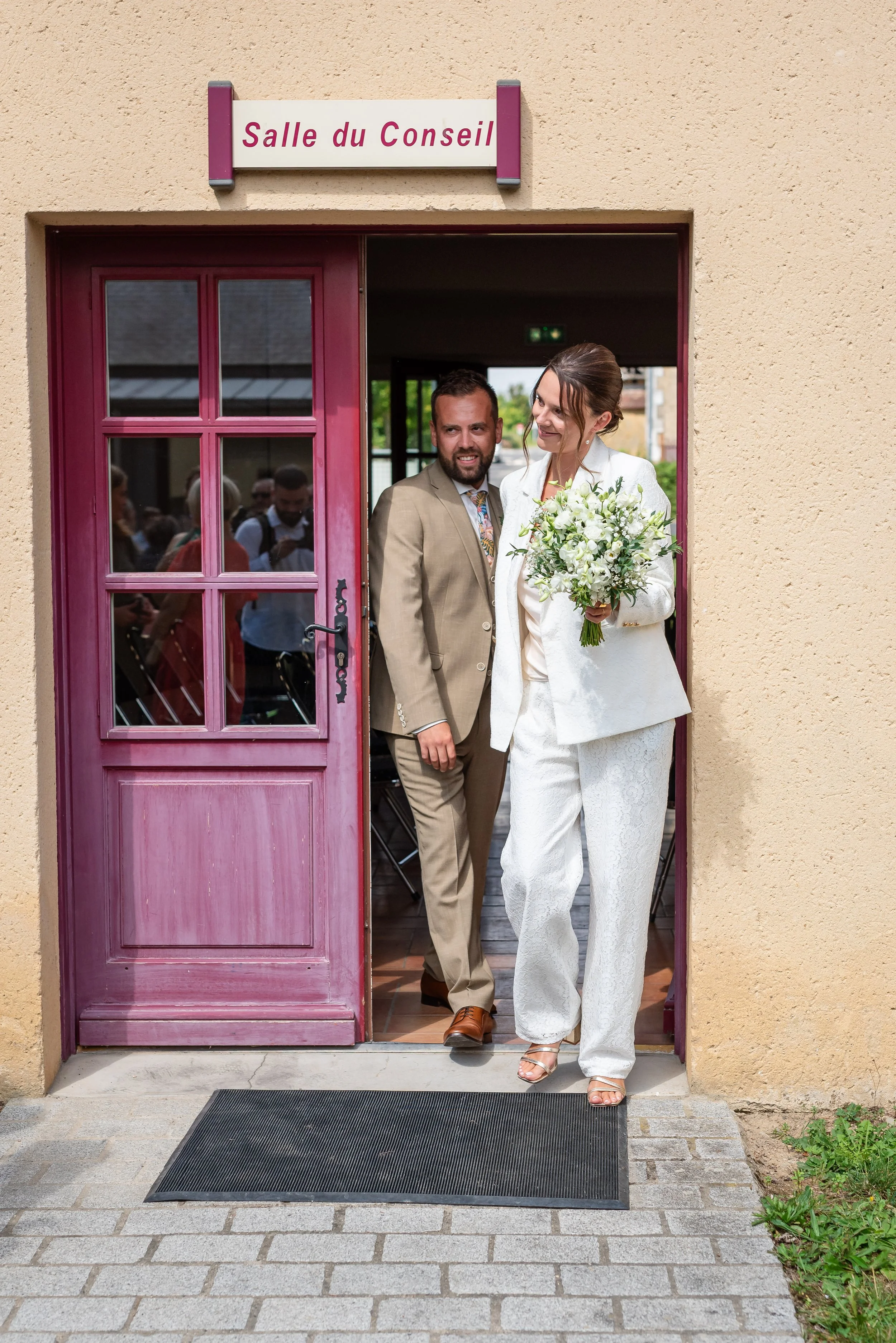 Un couple de mariés sortant d'une salle de réunion, la femme porte une robe blanche avec un bouquet de fleurs, l'homme porte un costume beige, ils sont souriants et visibles à travers la porte rose.