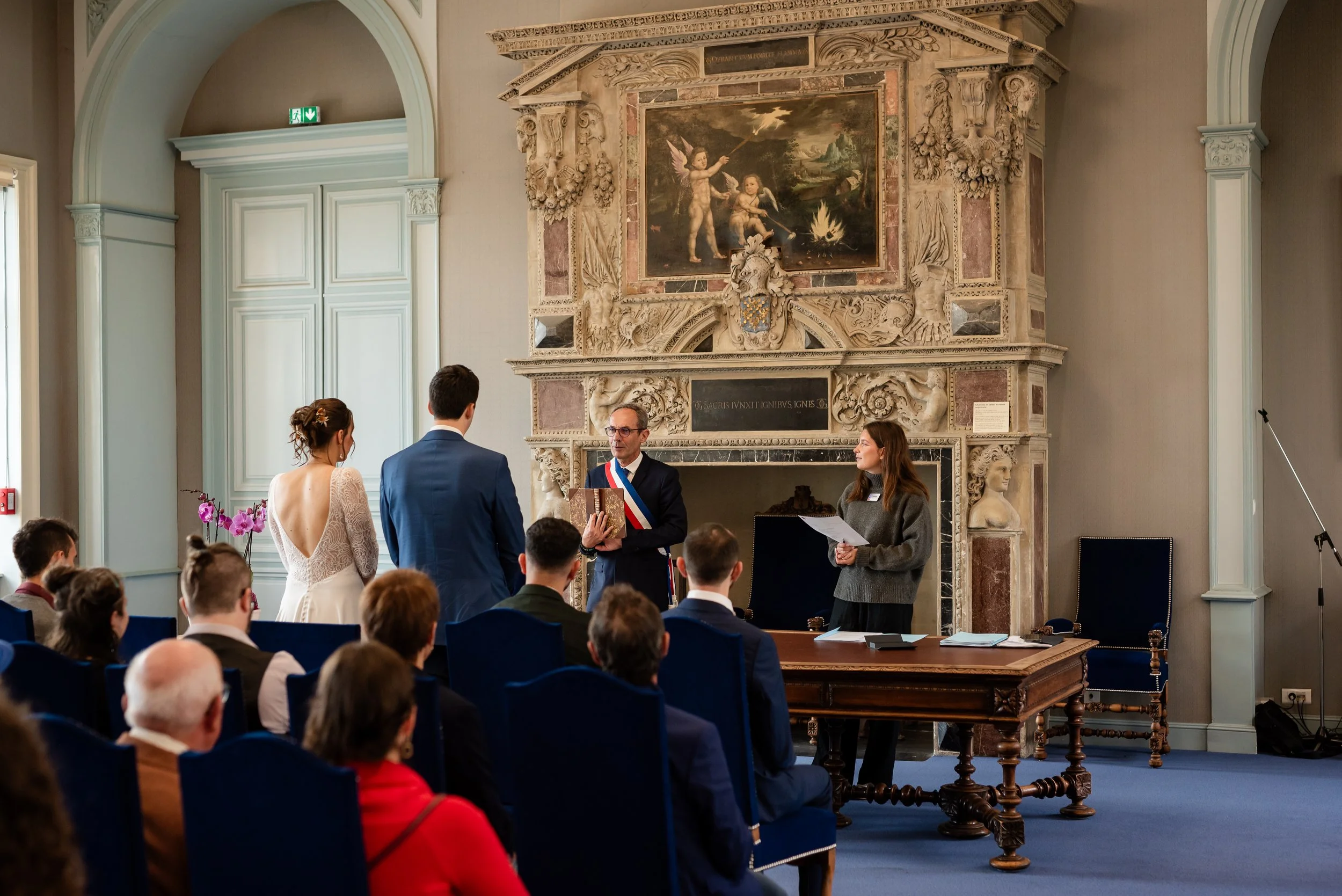 Une cérémonie officielle dans une salle ornée d'une cheminée en pierre et d'un tableau allégorique. Un homme avec un chapeau et un sash tricolore reçoit un diplôme ou un document d'une femme, tandis qu'une autre personne à côté, aussi avec un documen