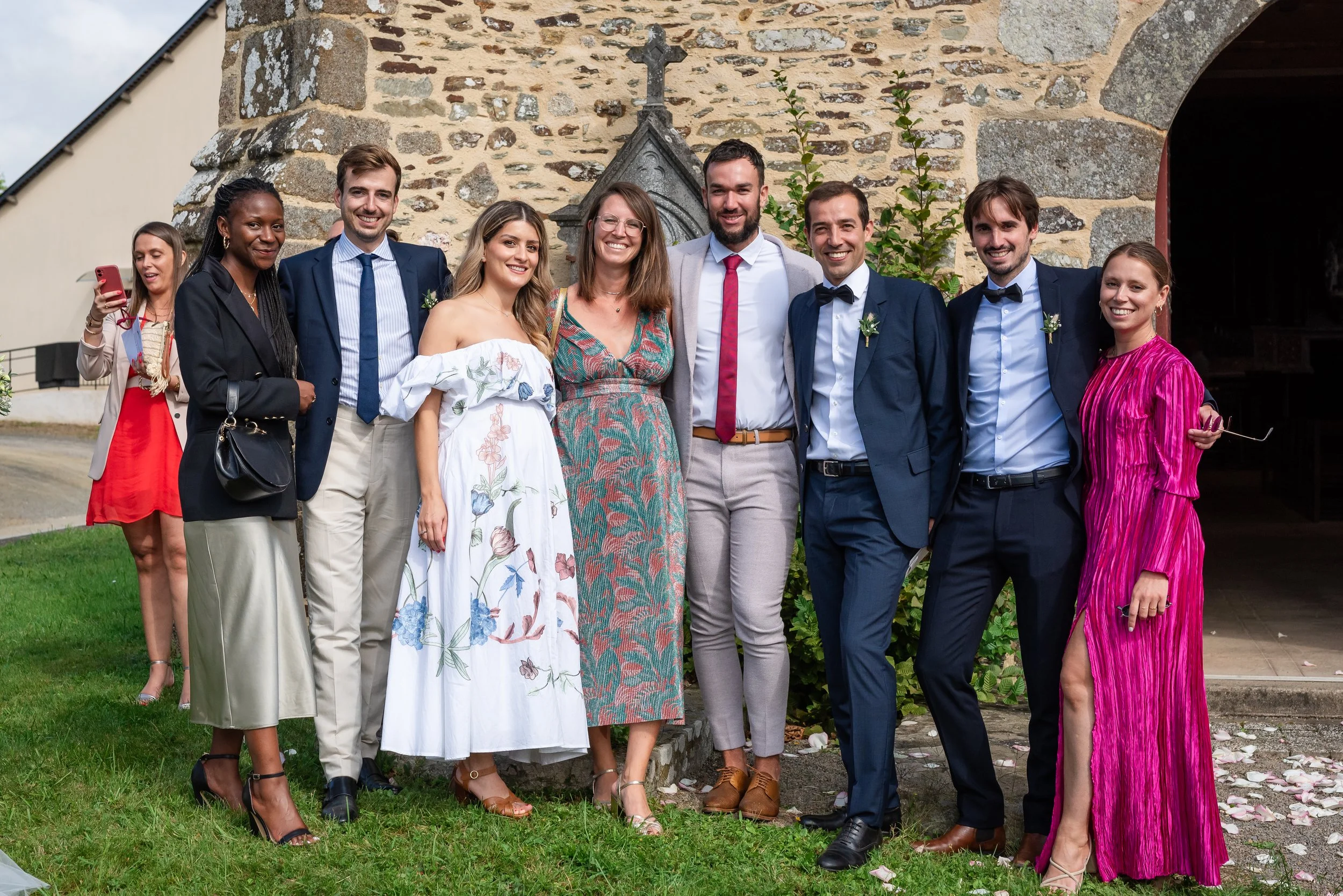 Un groupe de huit personnes posant pour une photo en extérieur devant un mur en pierre, lors d'une occasion spéciale ou célébration, avec un petit arbre en arrière-plan, certains en tenue formelle, d'autres en vêtements colorés.