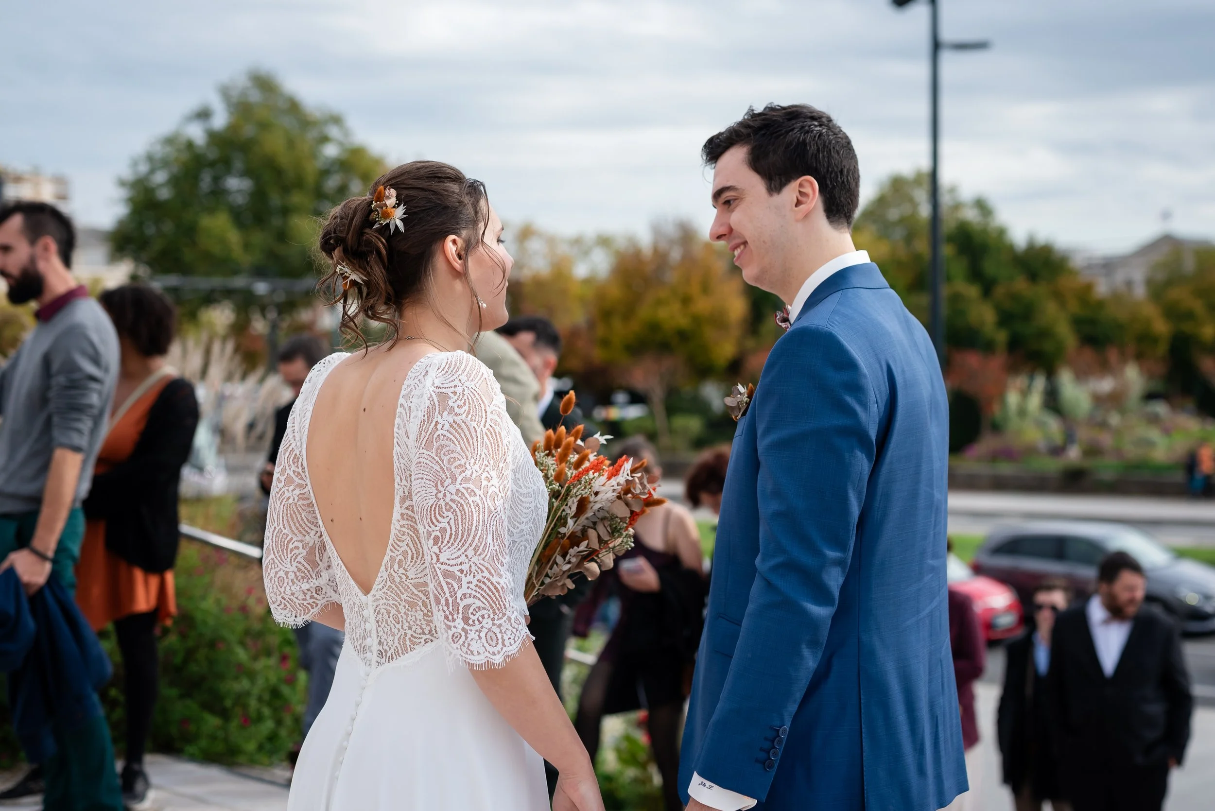 Un couple de mariés s'échangeant un sourire lors de leur mariage en plein air, avec des invités en arrière-plan et des arbres.