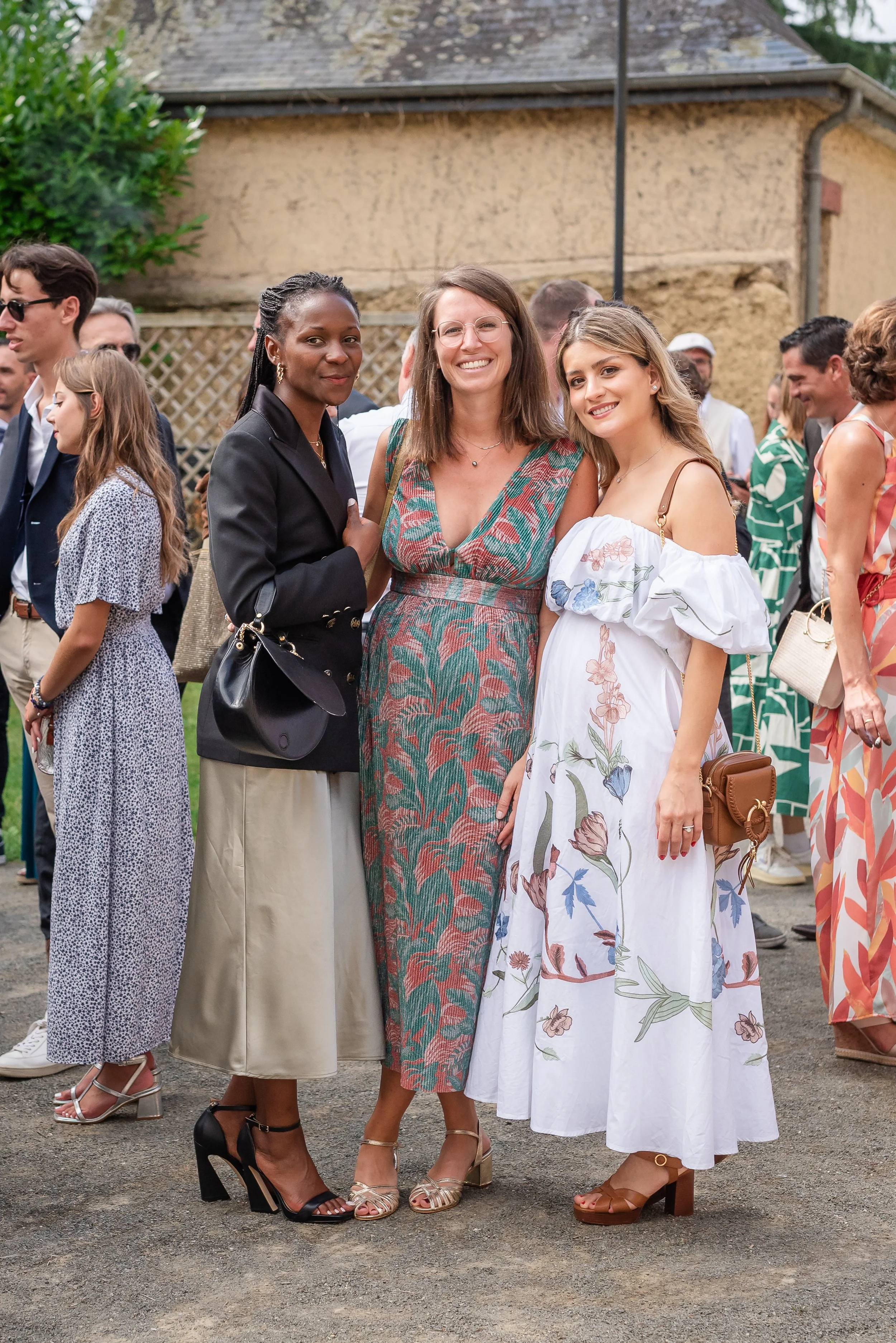 Trois femmes posent pour une photo lors d'un événement en plein air, parmi d'autres personnes en arrière-plan.