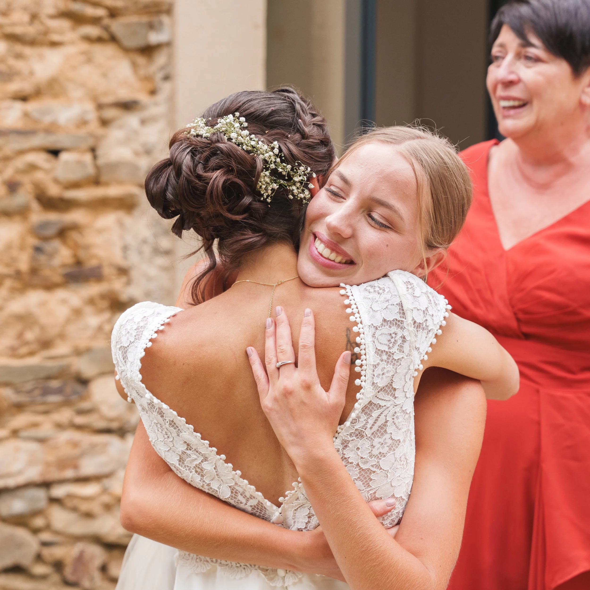 Deux femmes sont en train de partager un moment d'émotion, l'une porte une robe blanche avec des détails en dentelle et une couronne de fleurs dans les cheveux, l'autre porte un haut corail. Elles semblent se faire une accolade chaleureuse, avec un m