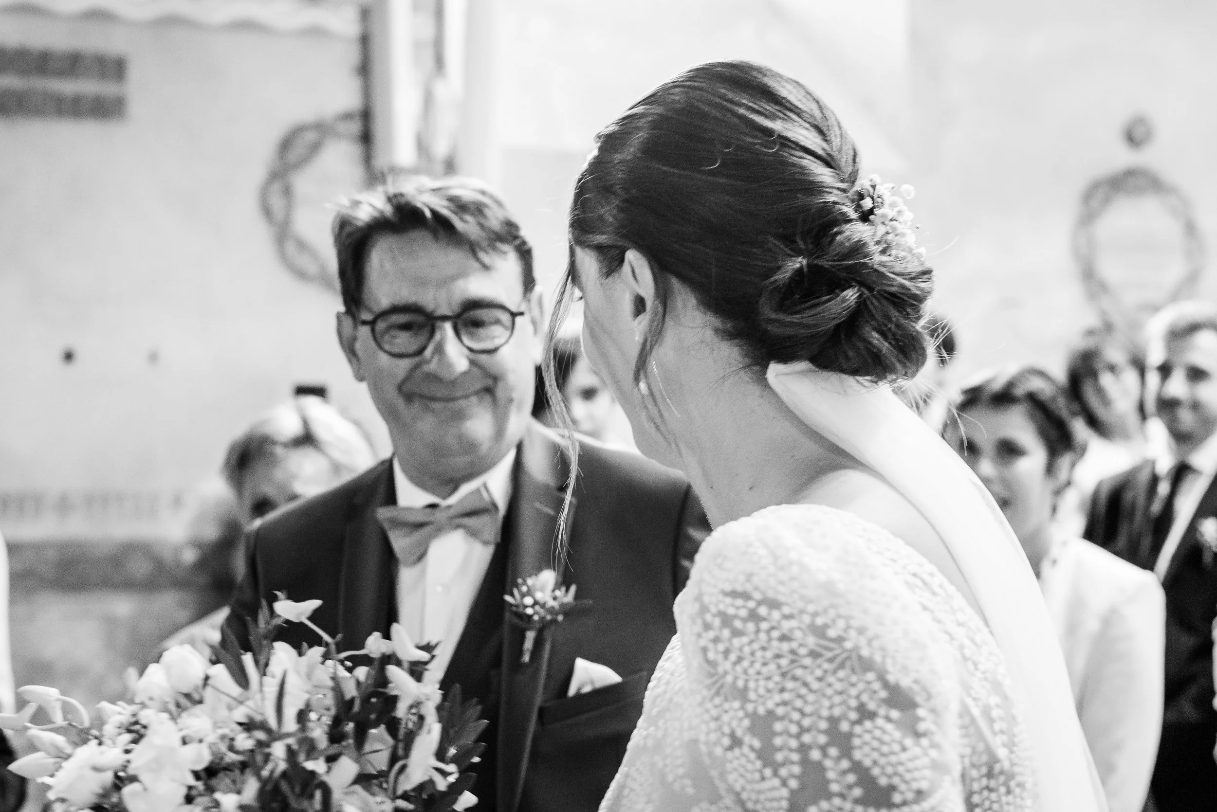 Un couple de mariés lors de leur mariage, avec le marié souriant et la mariée dont on voit le profil, dans une salle d'événement. Fond flou avec des invités.