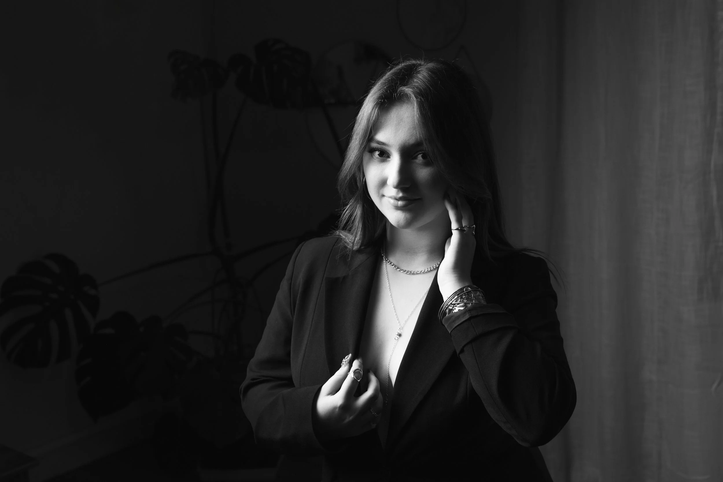 Une jeune femme avec des cheveux longs, portant une veste noire, des bijoux et posant devant un mur avec des plantes en arrière-plan, en noir et blanc.
