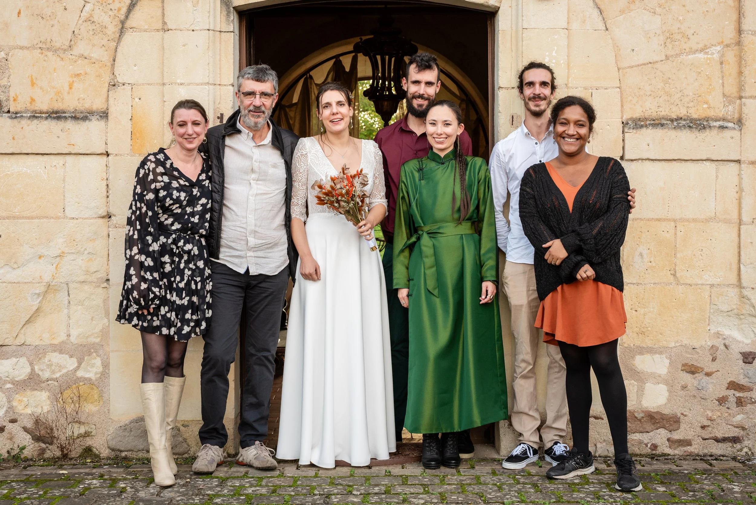 Groupe de sept personnes posant devant une porte en pierre, dont une femme en robe blanche tenant un bouquet de fleurs, vraisemblablement à l'occasion d'une célébration ou cérémonie.