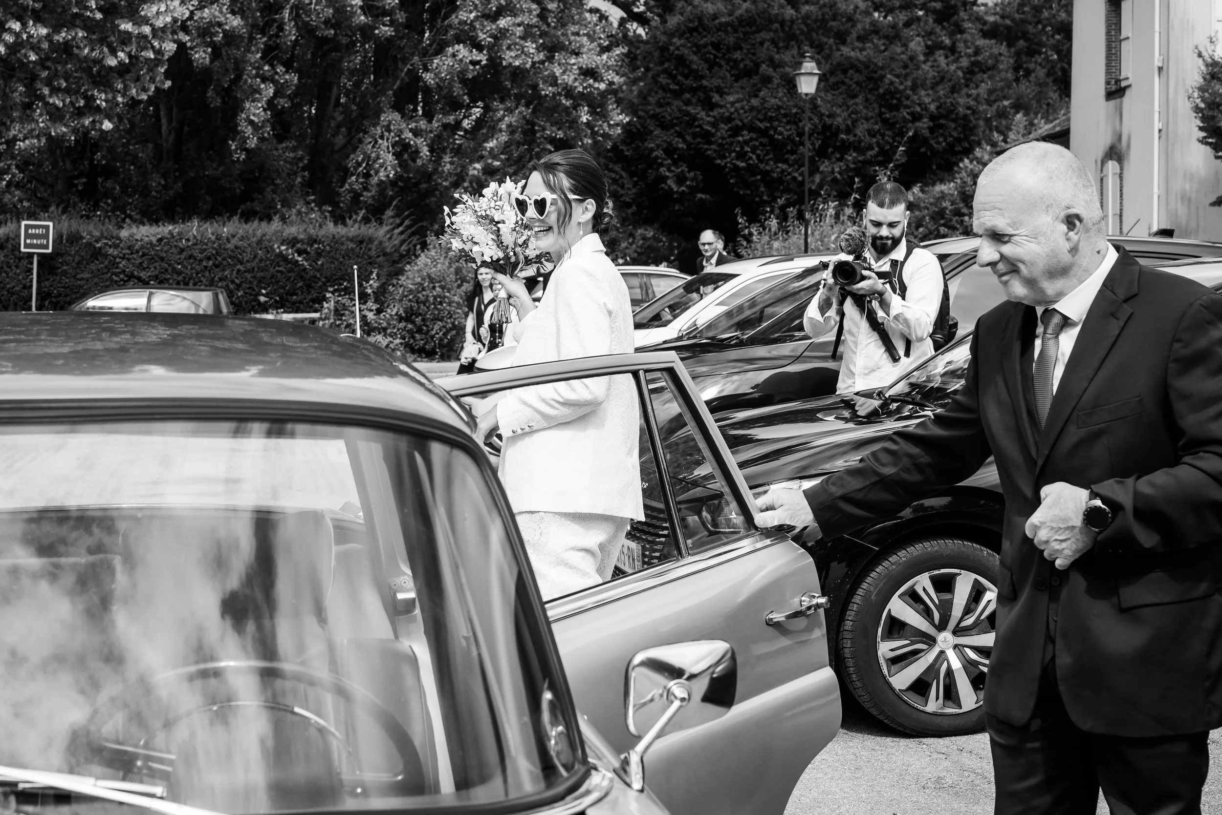 Une femme avec des lunettes en forme de cœurs, tenant un bouquet de fleurs, souriant à côté d'une voiture, en arrière-plan des personnes avec un appareil photo, dans une scène de mariage en extérieur.