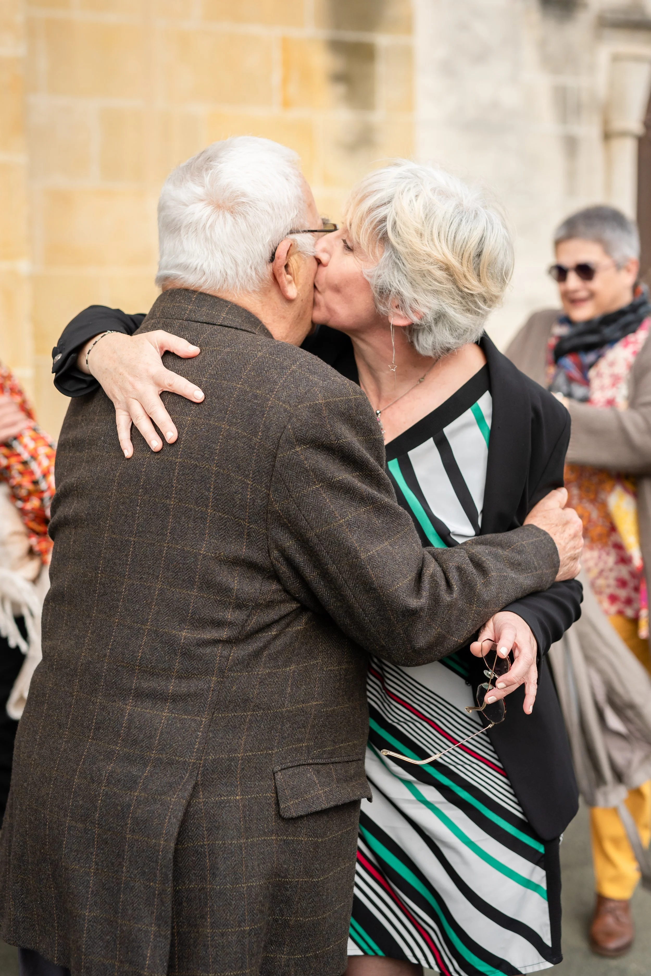 Une femme et un homme âgés s'embrassent en se tenant dans une embrassade chaleureuse lors d'une célébration. La femme porte une robe rayée et la femme un manteau à carreaux. En arrière-plan, d'autres personnes sont présentes, dont une femme avec des 