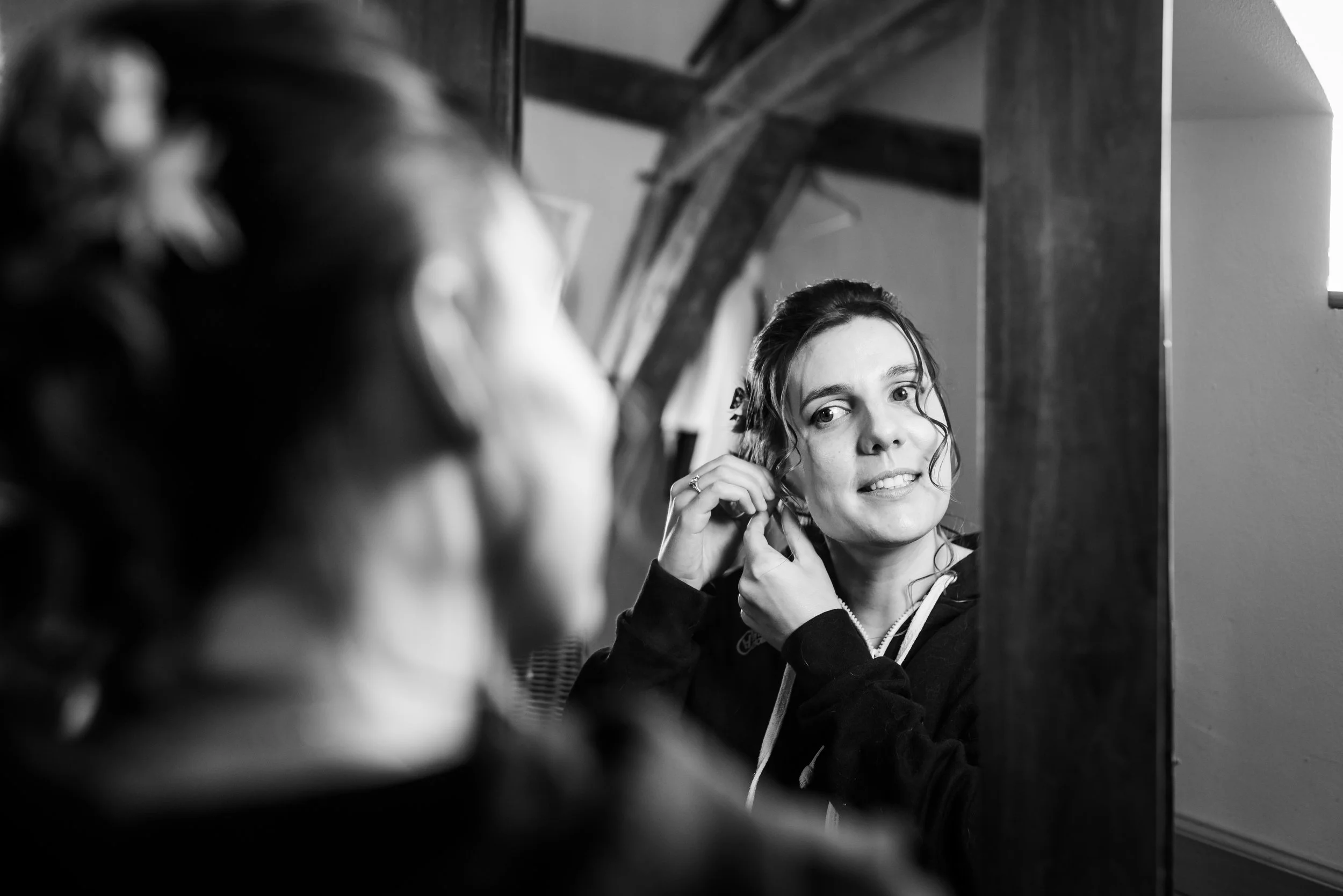 Une femme se regarde dans un miroir, ajustant ses boucles de cheveux, avec un sourire léger. La photo est en noir et blanc et prise dans une pièce avec une structure en bois visible en arrière-plan.
