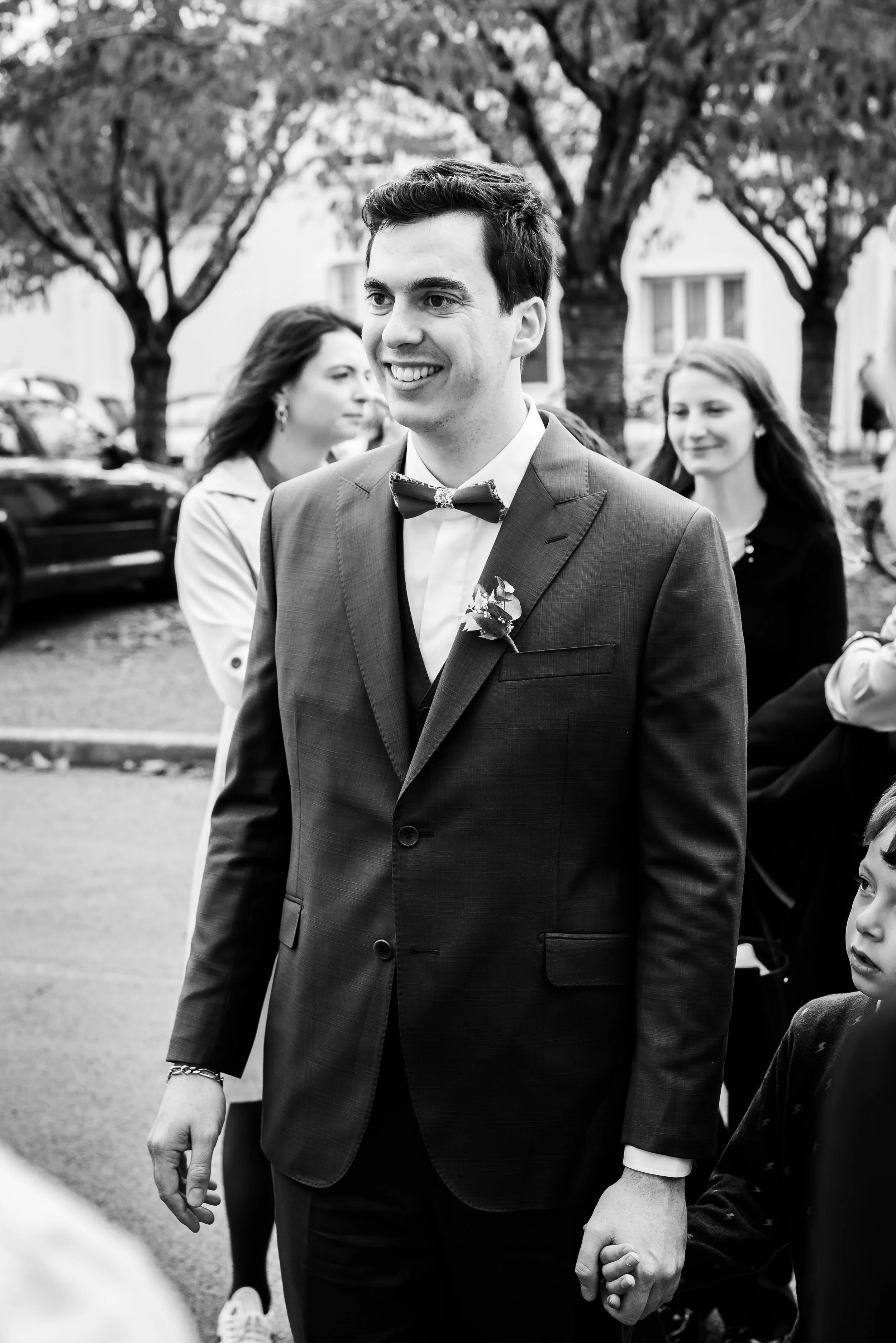 Un homme en costume et nœud papillon, souriant, tenant la main d'un enfant lors d'une cérémonie ou d'un événement en plein air, avec des personnes et des arbres en arrière-plan.