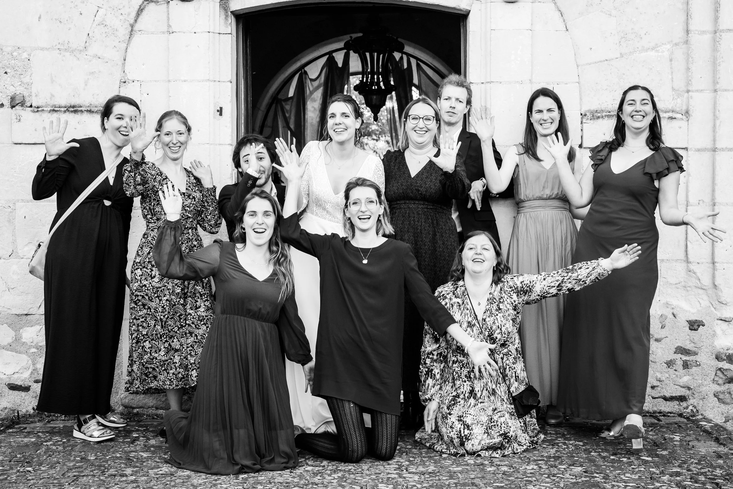 Un groupe de dix personnes souriantes et levant la main devant une façade en pierre, posant pour une photo de groupe en plein air.