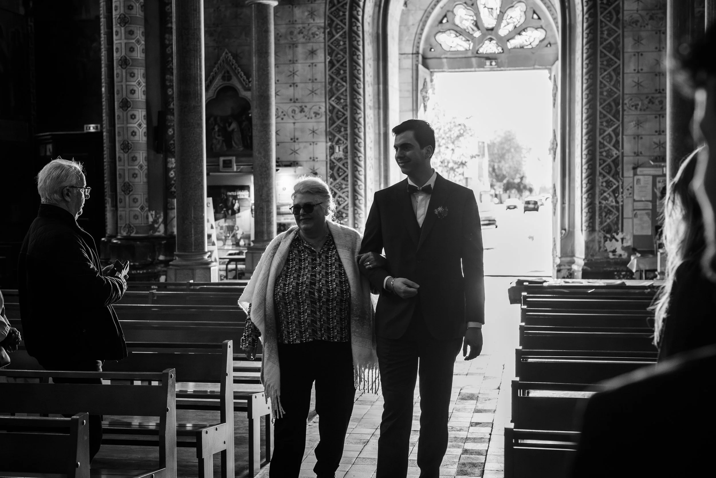 Une scène dans une église avec plusieurs personnes, dont un homme en costume avec un nœud papillon, une femme portant des lunettes et un foulard, et une personne âgée avec des lunettes, à l'intérieur d'une église aux murs ornés et fenêtres colorées.