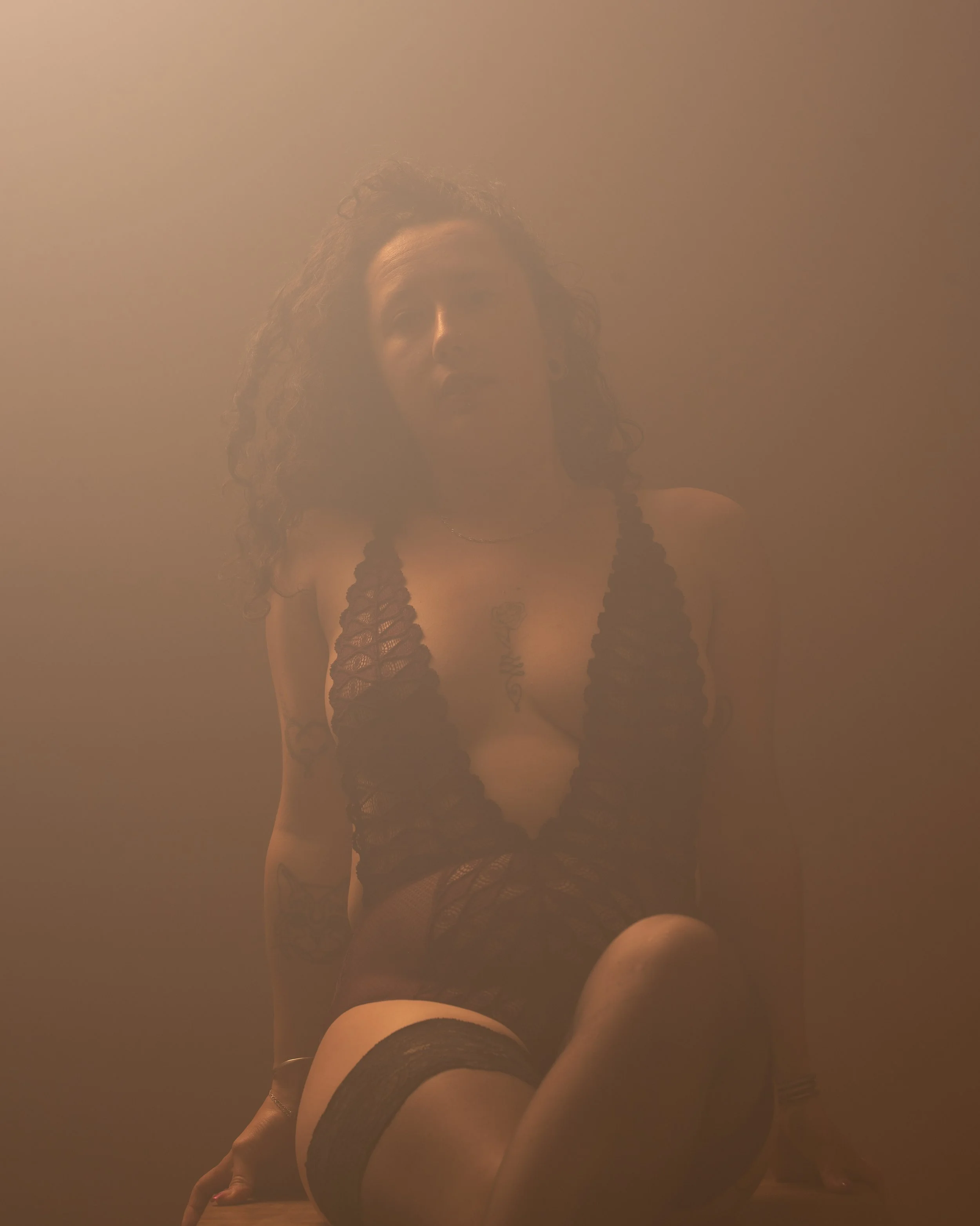 Une femme en lingerie noire et collants noirs, vue sous un angle basses, posant contre un mur beige, avec un peu de fumée ou brouillard autour.