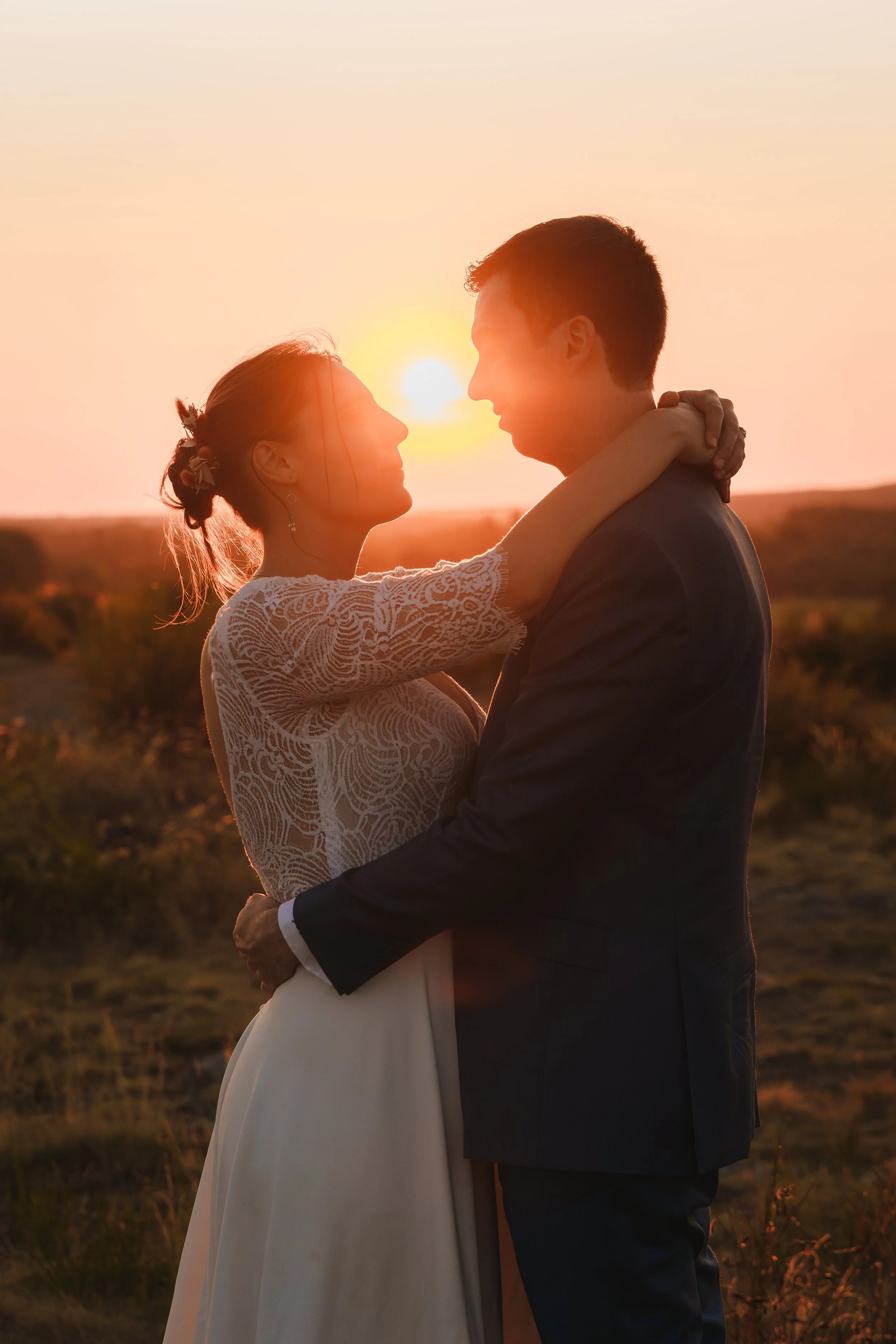 Un couple qui s'enlace tendrement au coucher du soleil dans un champ, la femme porte une robe blanche en dentelle et le homme un costume sombre.