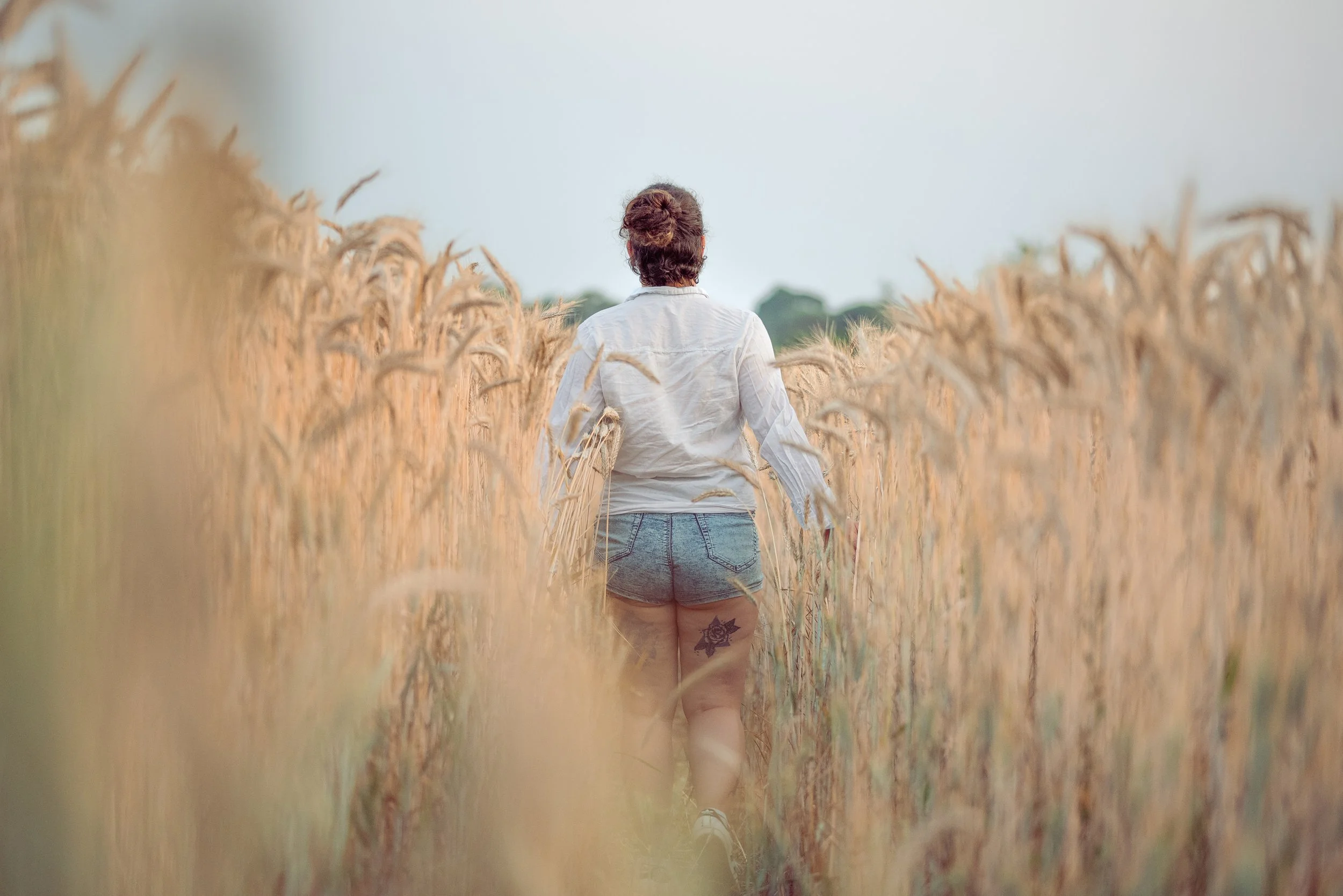 Une femme marche dans un champ de blé doré, vue de dos, portant un chemisier blanc et un short en denim, avec un tatouage de rose sur la cuisse. La scène se déroule en plein jour, avec un ciel partiellement nuageux.