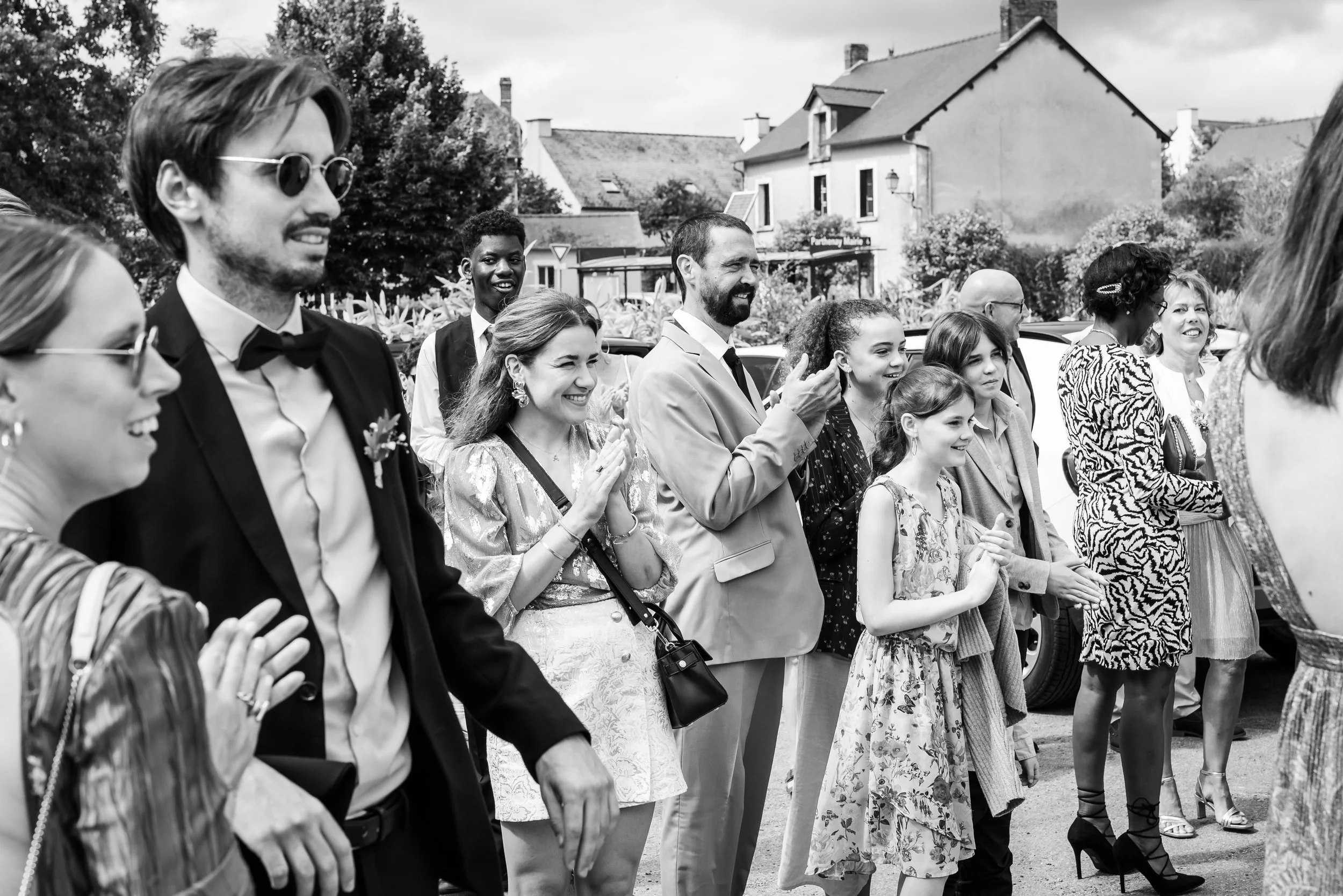 Un groupe de personnes assises ou debout lors d'un événement extérieur, souriant et applaudissant, dans un cadre urbain avec des bâtiments en arrière-plan.