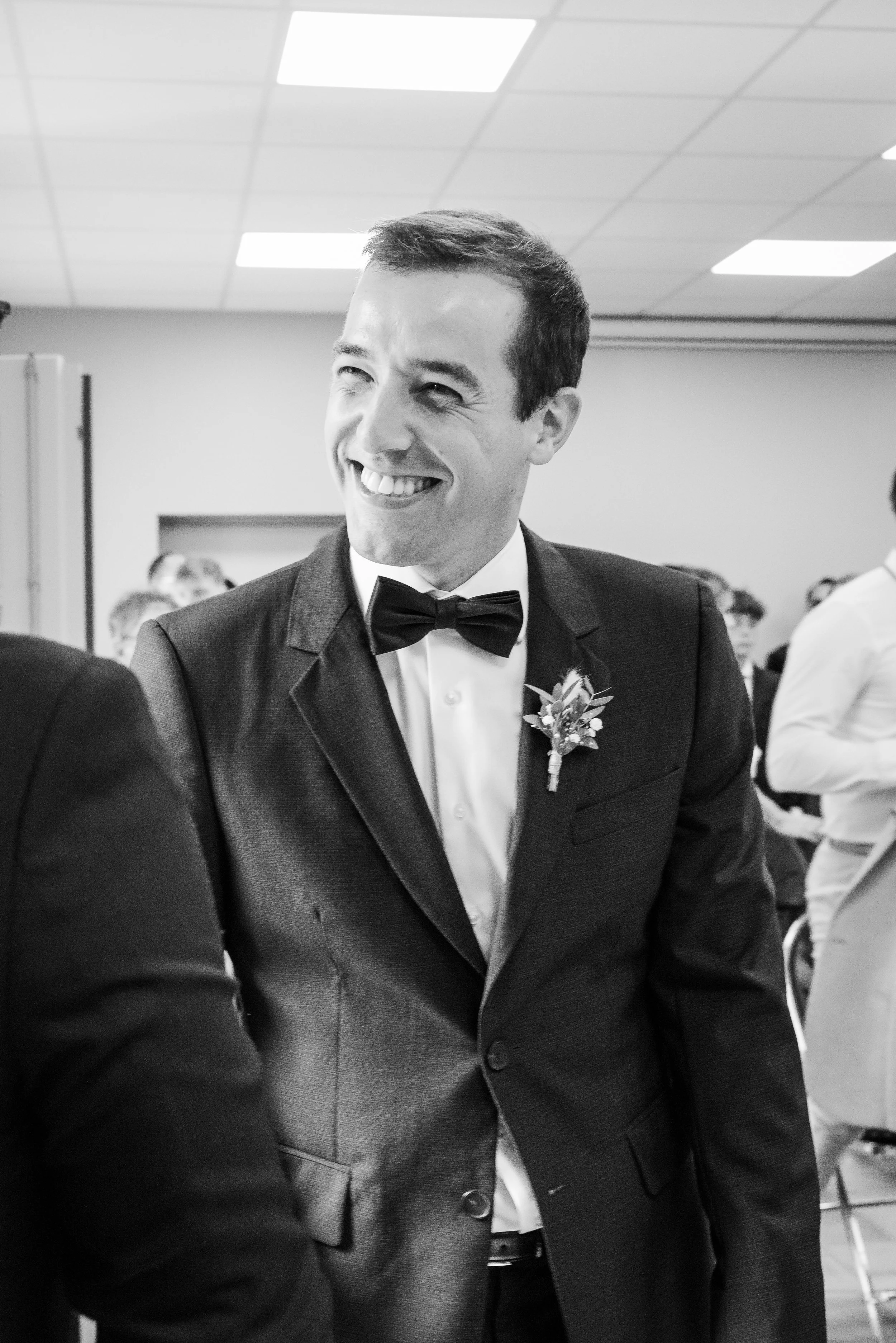 Homme souriant en costume noir et nœud papillon lors d'un événement formel, avec d'autres personnes en arrière-plan dans une salle basse lumière avec plafond en dalles.