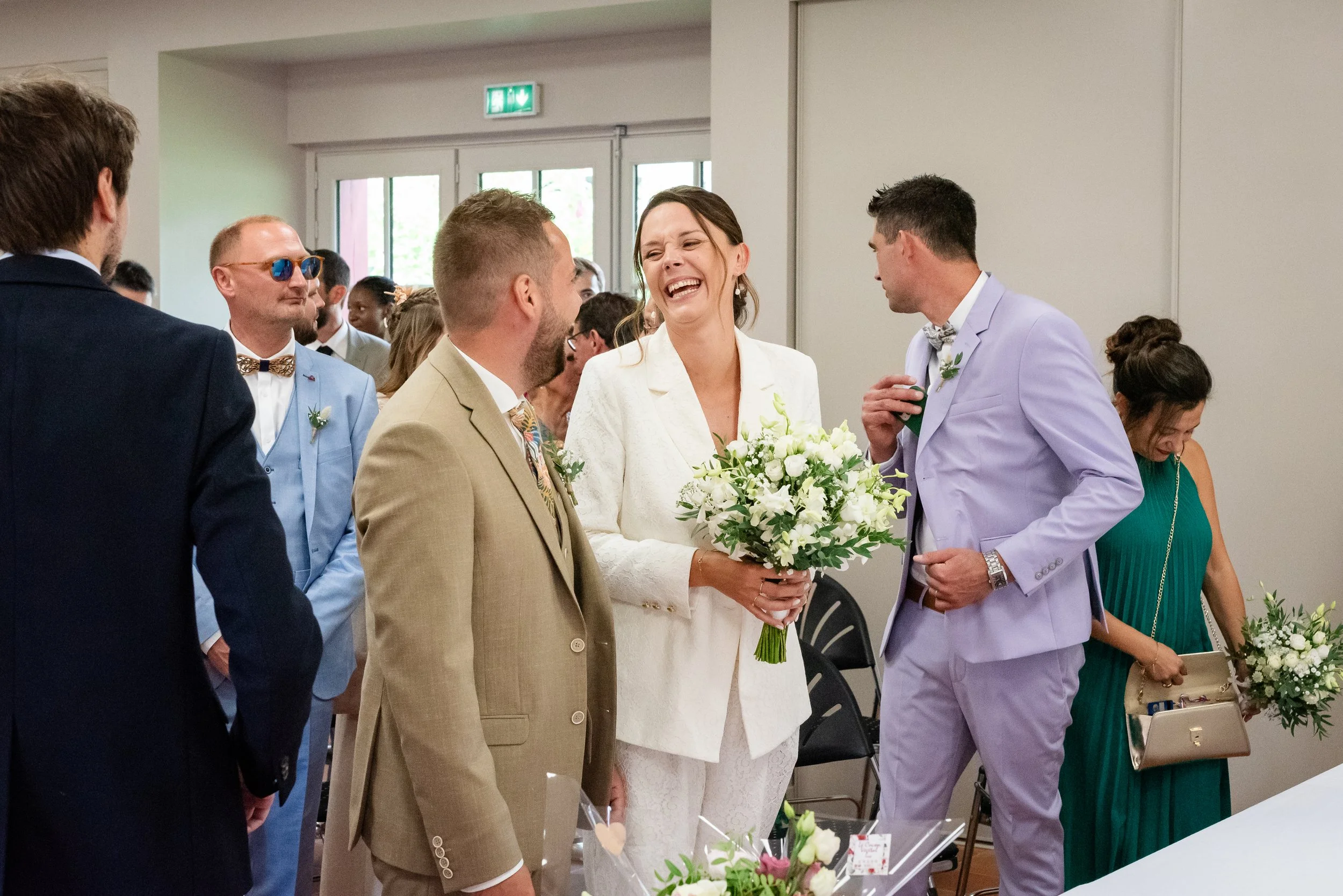 Un groupe de personnes habillées élégamment, probablement lors d'un mariage ou d'une cérémonie, discutent et rient dans une salle intérieure.