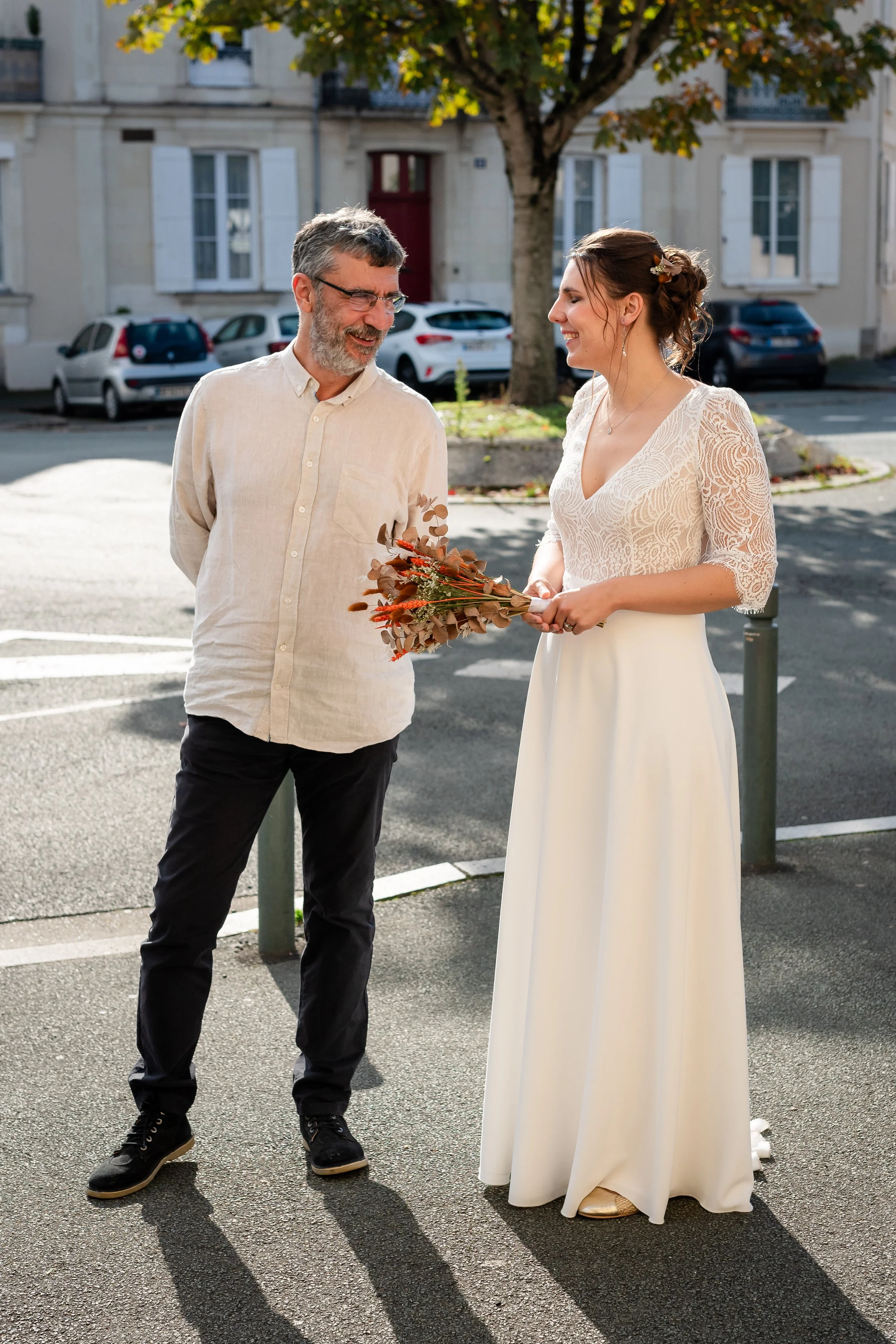 Un homme et une femme souriant dehors, avec la femme tenant un bouquet de fleurs, à côté d'un arbre et de voitures en arrière-plan.