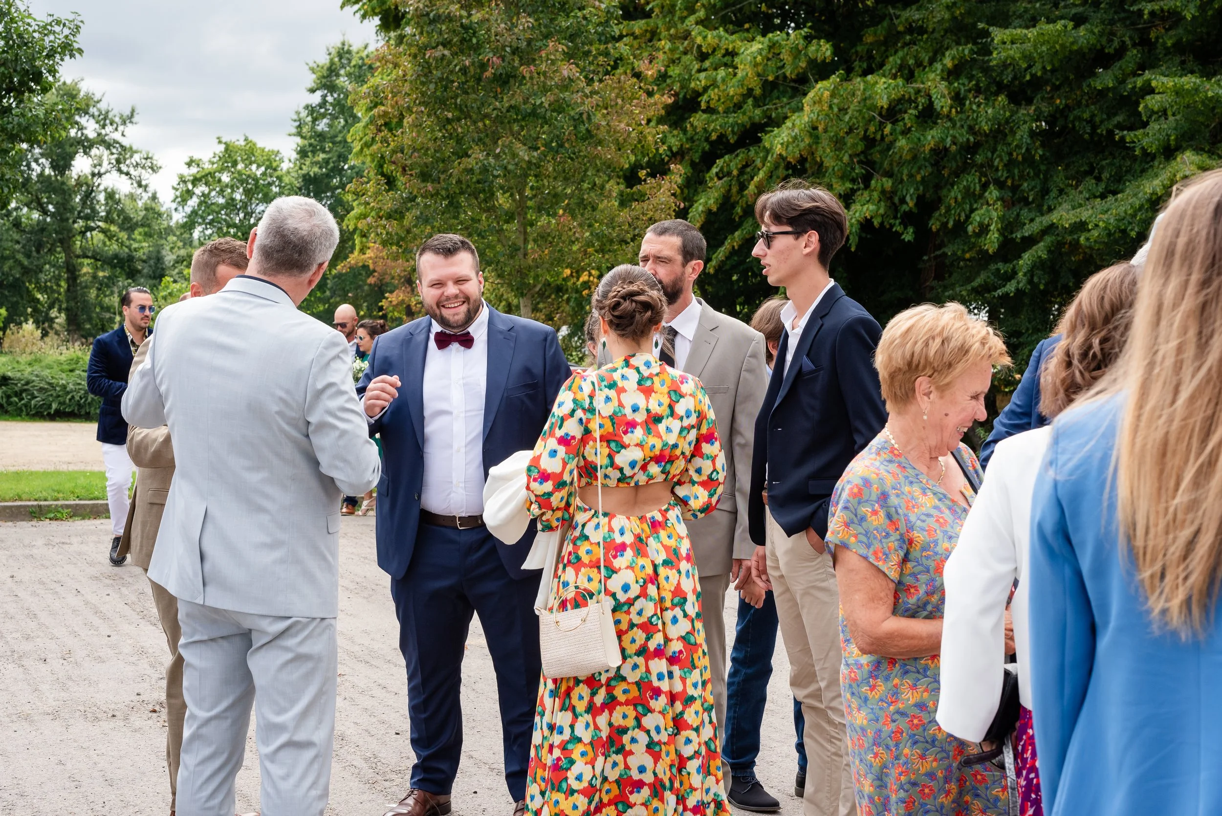Un groupe de personnes habillées élégamment, semblant participer à un événement social ou un mariage, discutant dehors sur fond d'arbres verts.
