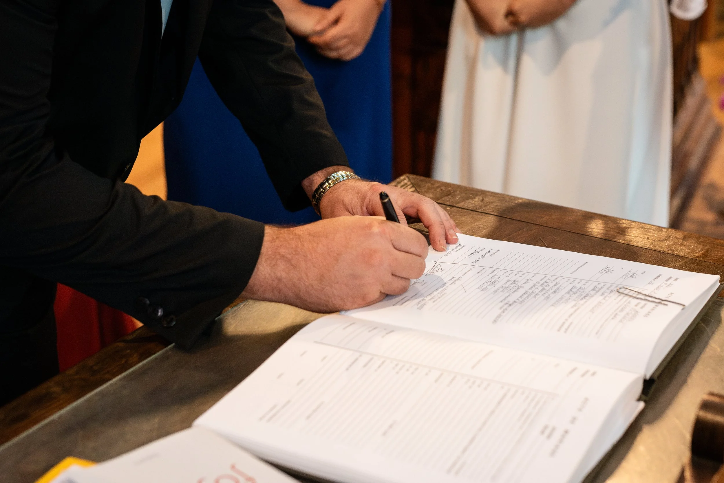 Une personne en costume noir signe un document sur une table en bois, avec d'autres personnes en arrière-plan.