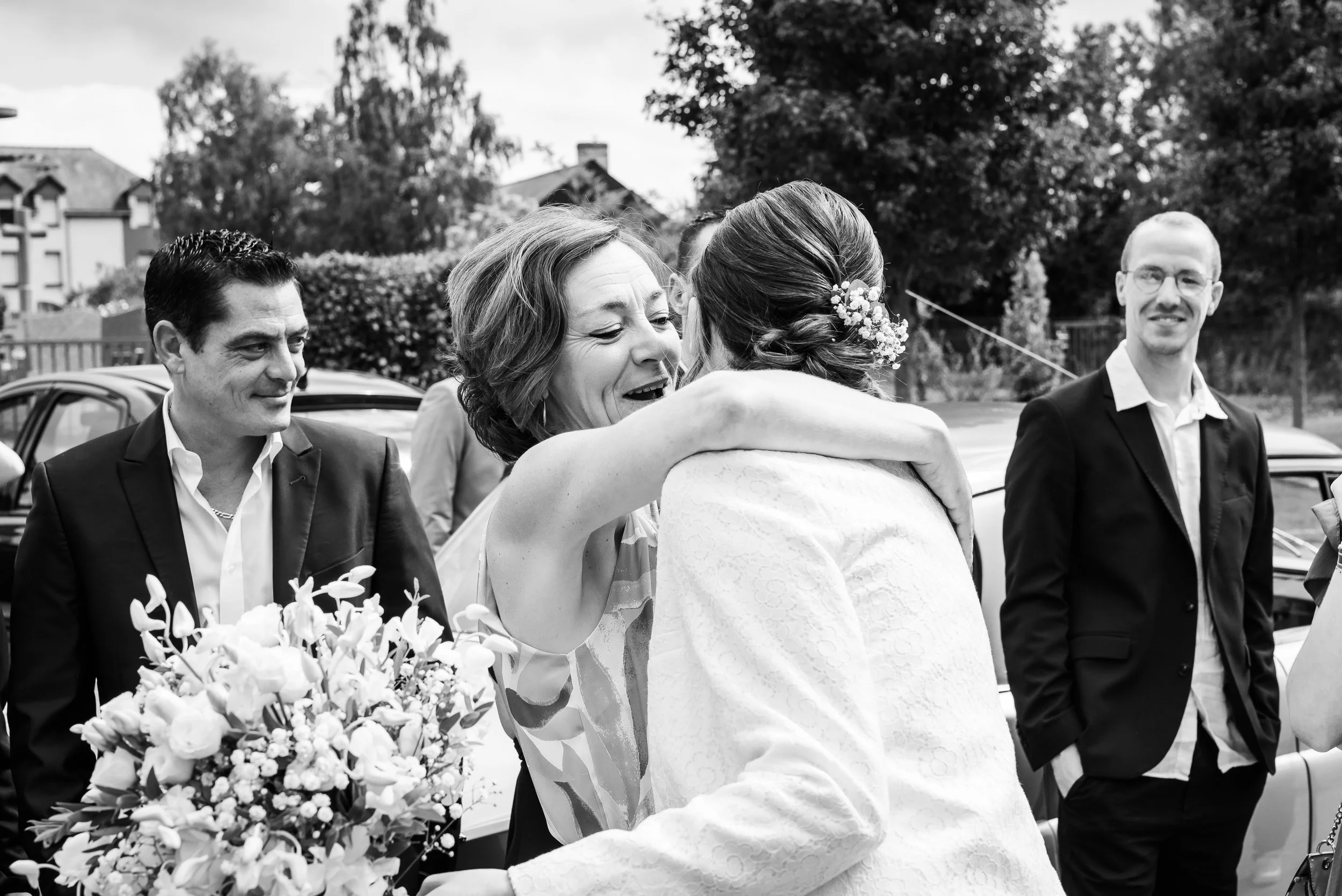 Groupe de personnes lors d'une cérémonie, femme en train d'embrasser une autre personne, bouquet de fleurs visible.