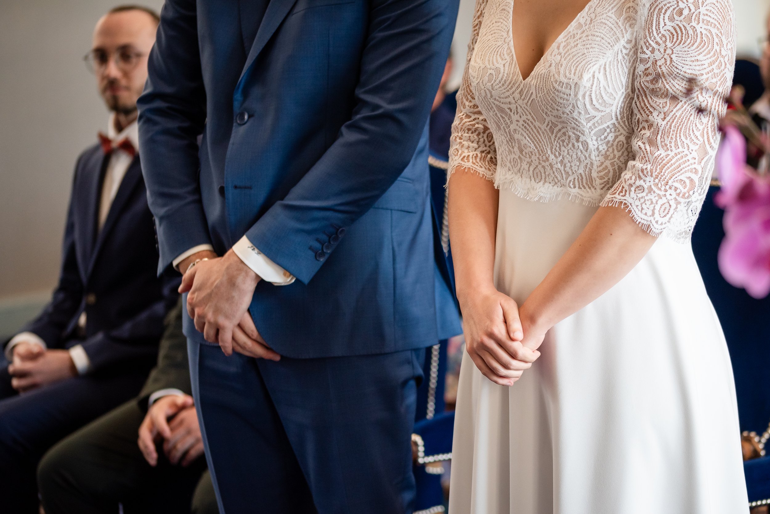 Gens en costume et robe de mariage se tenant par la main lors d'une cérémonie, avec d'autres personnes assises en arrière-plan.