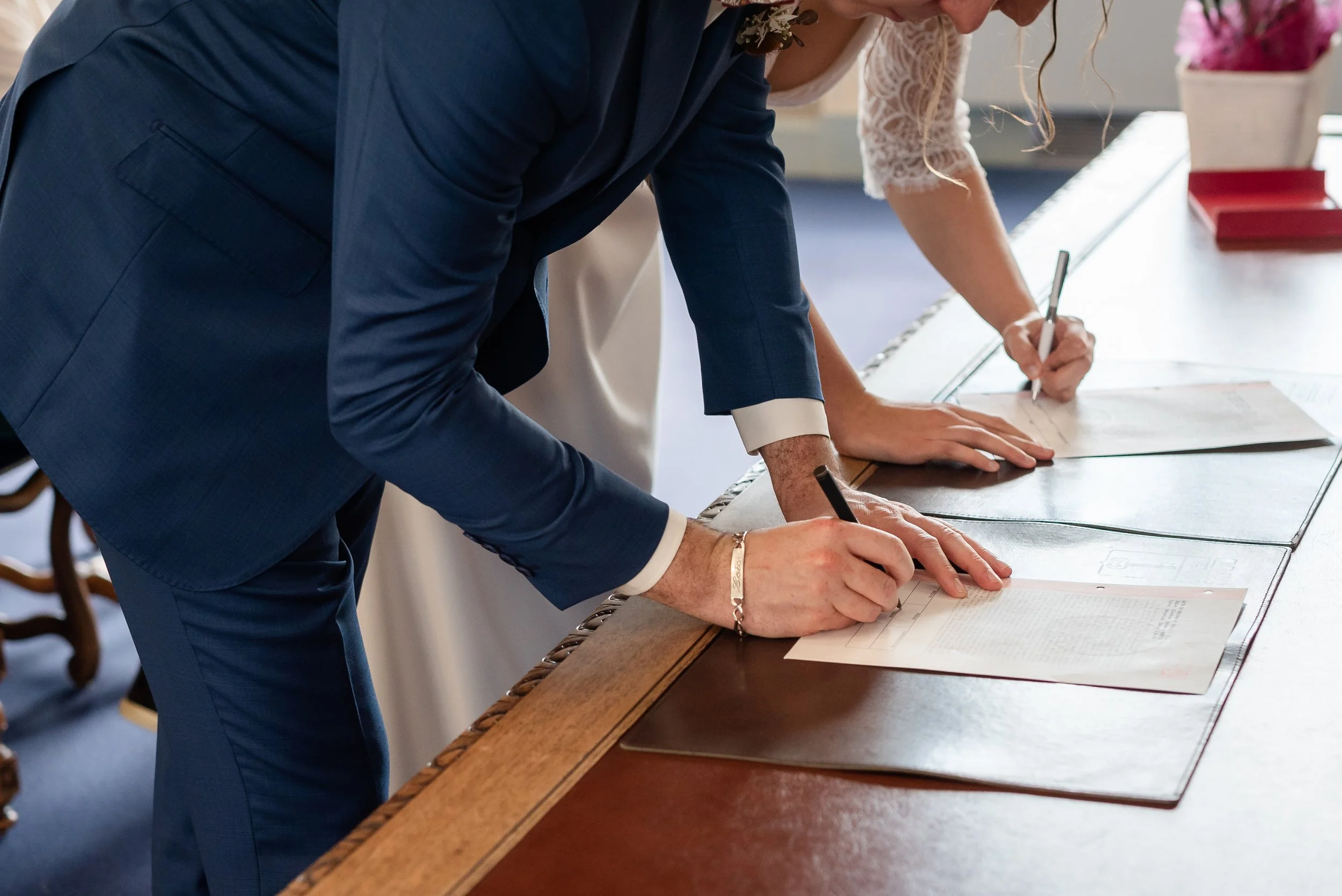 Deux personnes sont debout devant une table en train de signer des documents, l'une en costume bleu foncé et l'autre en vêtements blancs avec des manches en dentelle, dans une pièce éclairée naturellement.