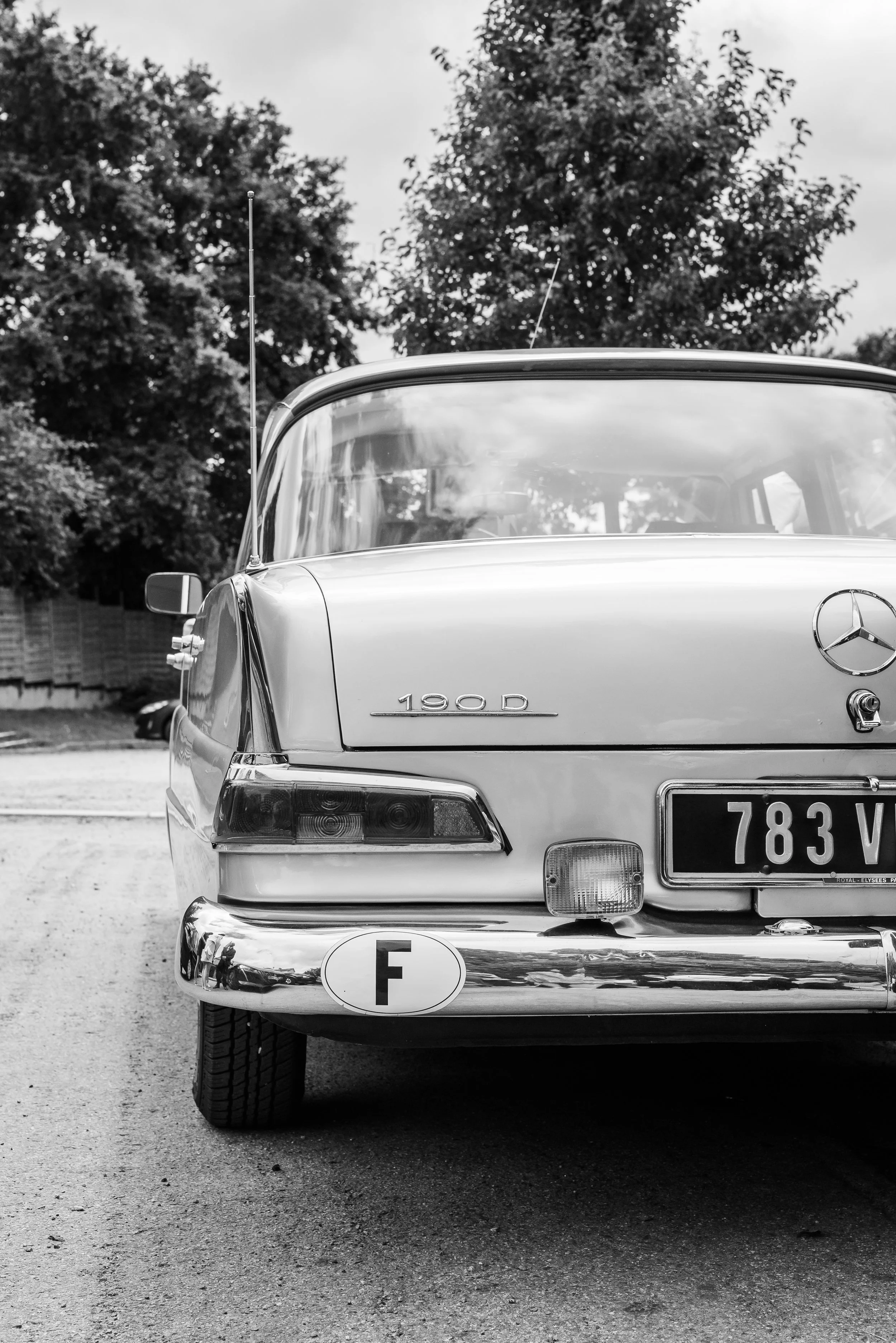 Voiture Mercedes-Benz classique noire vue de face, avec le badge et la plaque d'immatriculation visible, stationnée sur une route, avec des arbres en arrière-plan, en photographie en noir et blanc.
