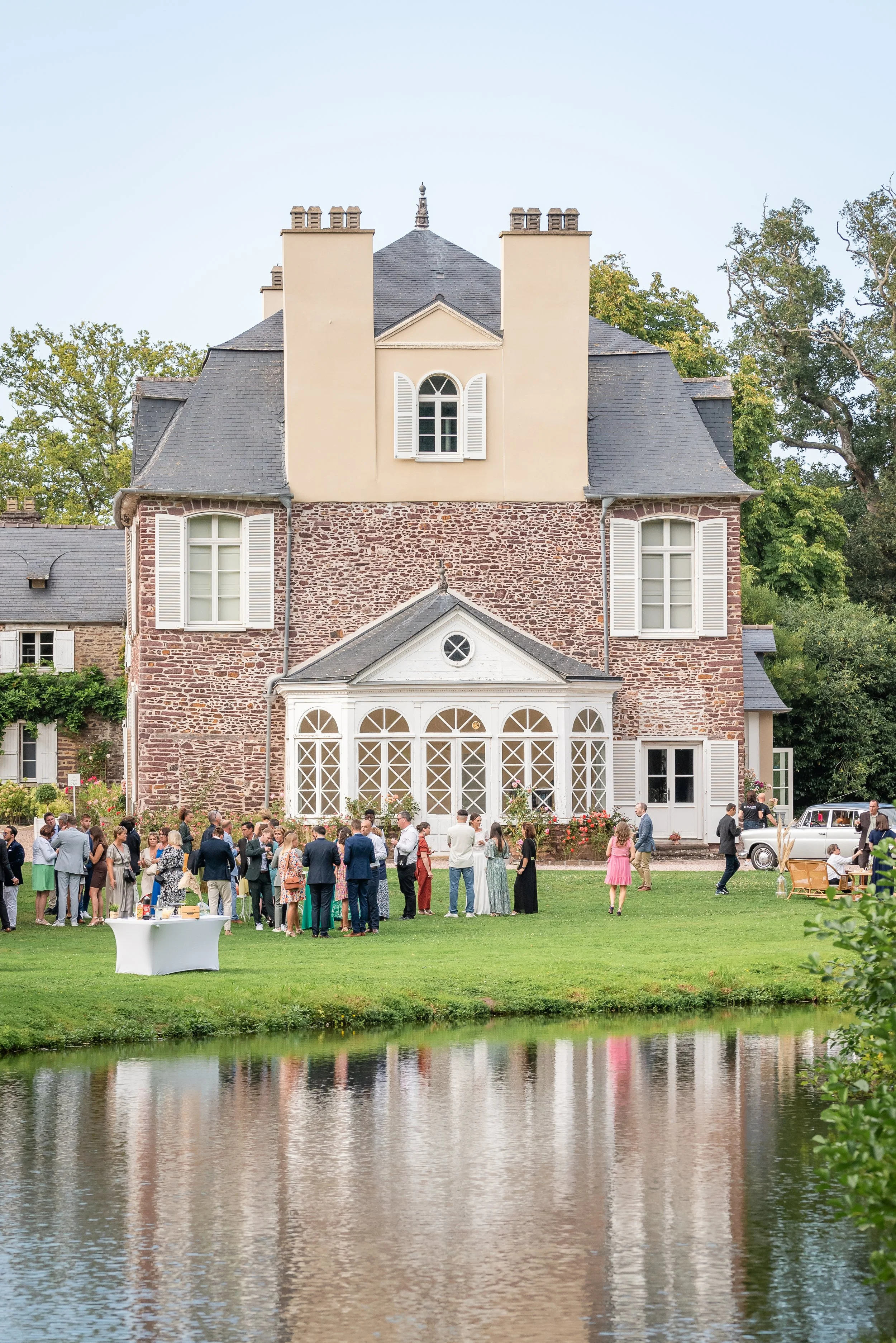 Une fête en plein air devant une grande maison en pierre avec un toit en ardoise et plusieurs fenêtres à volets blancs, des invités en costume et en robe discutent et s'amusent dans un jardin bien entretenu, près d'un lac, par une journée ensoleillée.