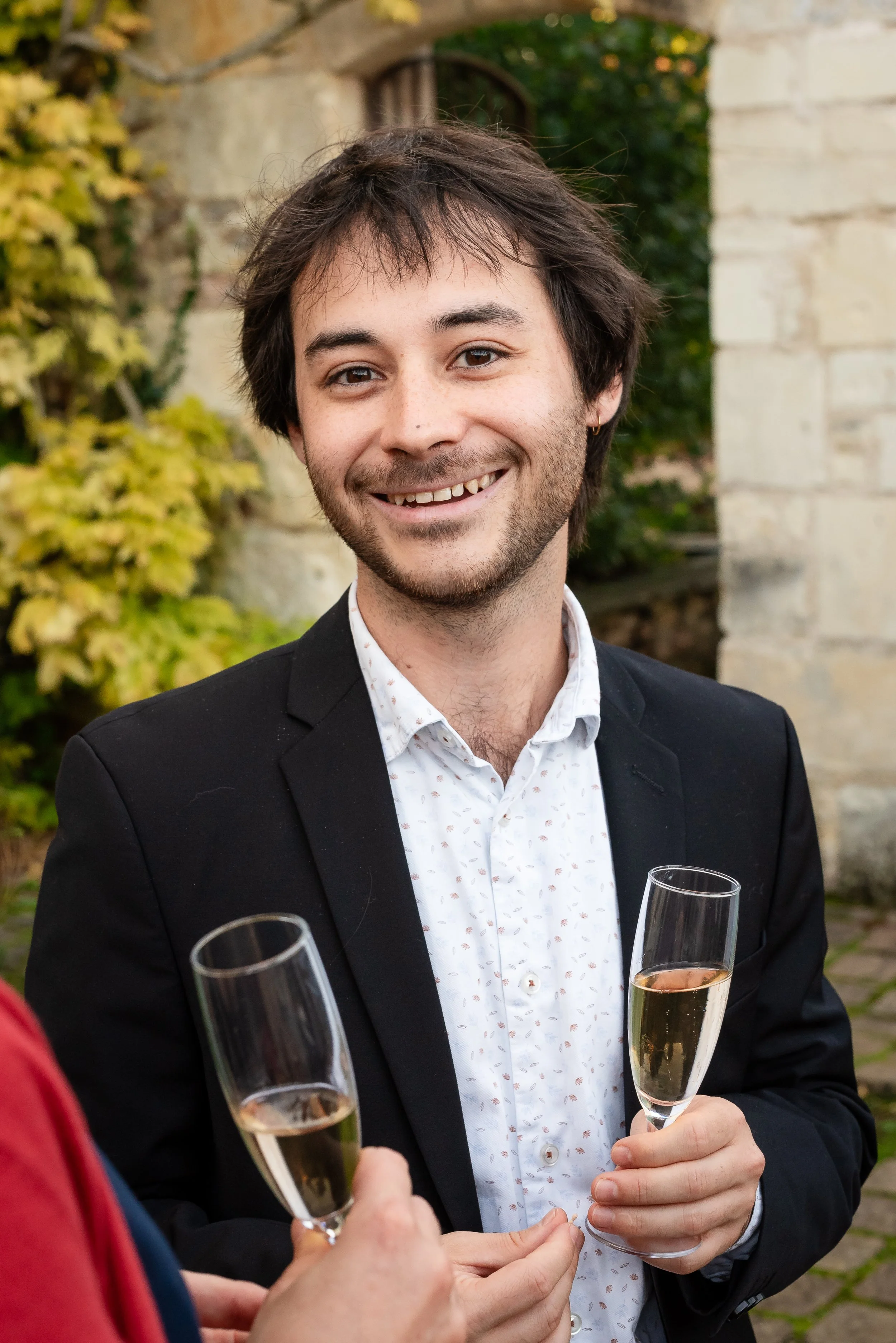 Deux personnes tiennent des verres de champagne lors d'une célébration en extérieur. Une personne porte un costume noir et une chemise blanche à motifs, souriant et regardant l'objectif, avec un fond de végétation et de pierre.
