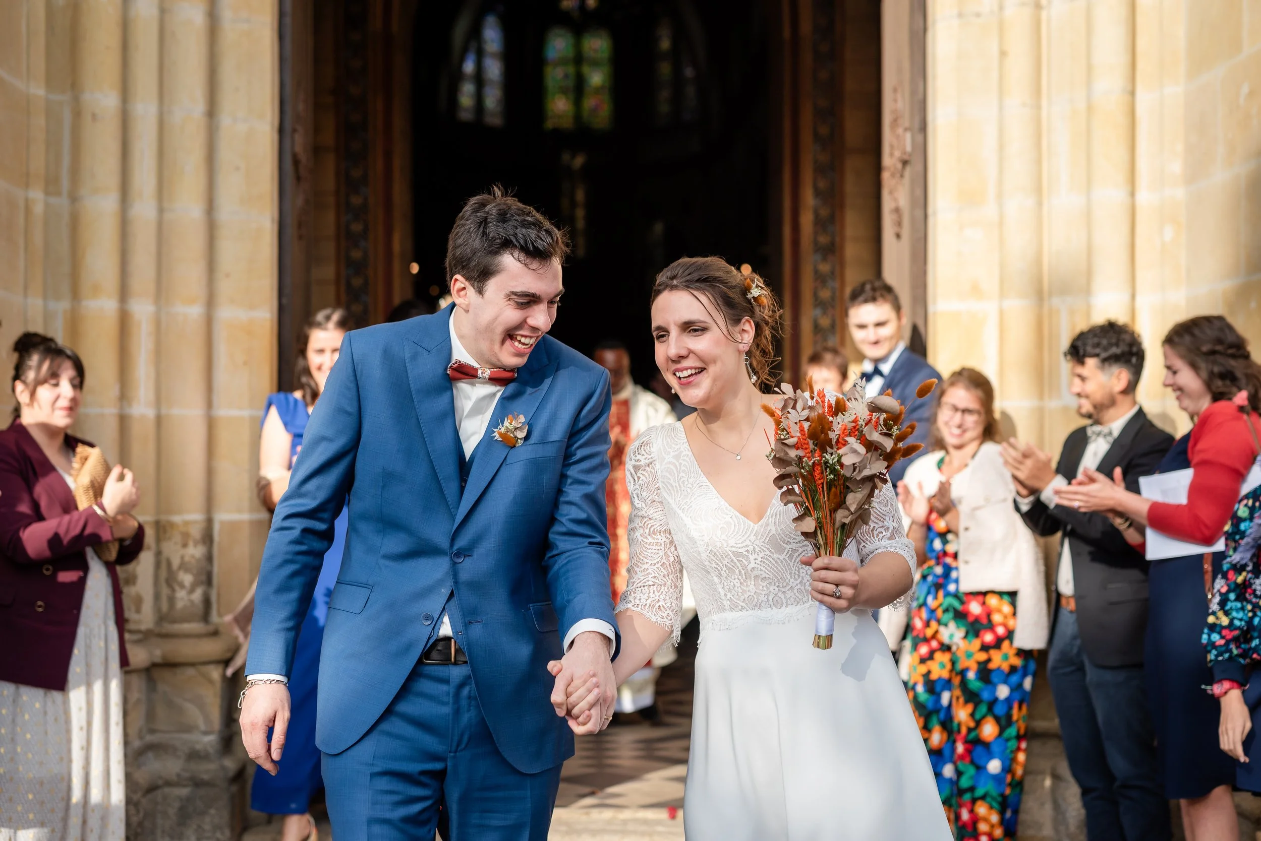 Un couple de mariés souriants se tenant la main à l'entrée d'une église, entourés d'invités applaudissant, lors de leur mariage.