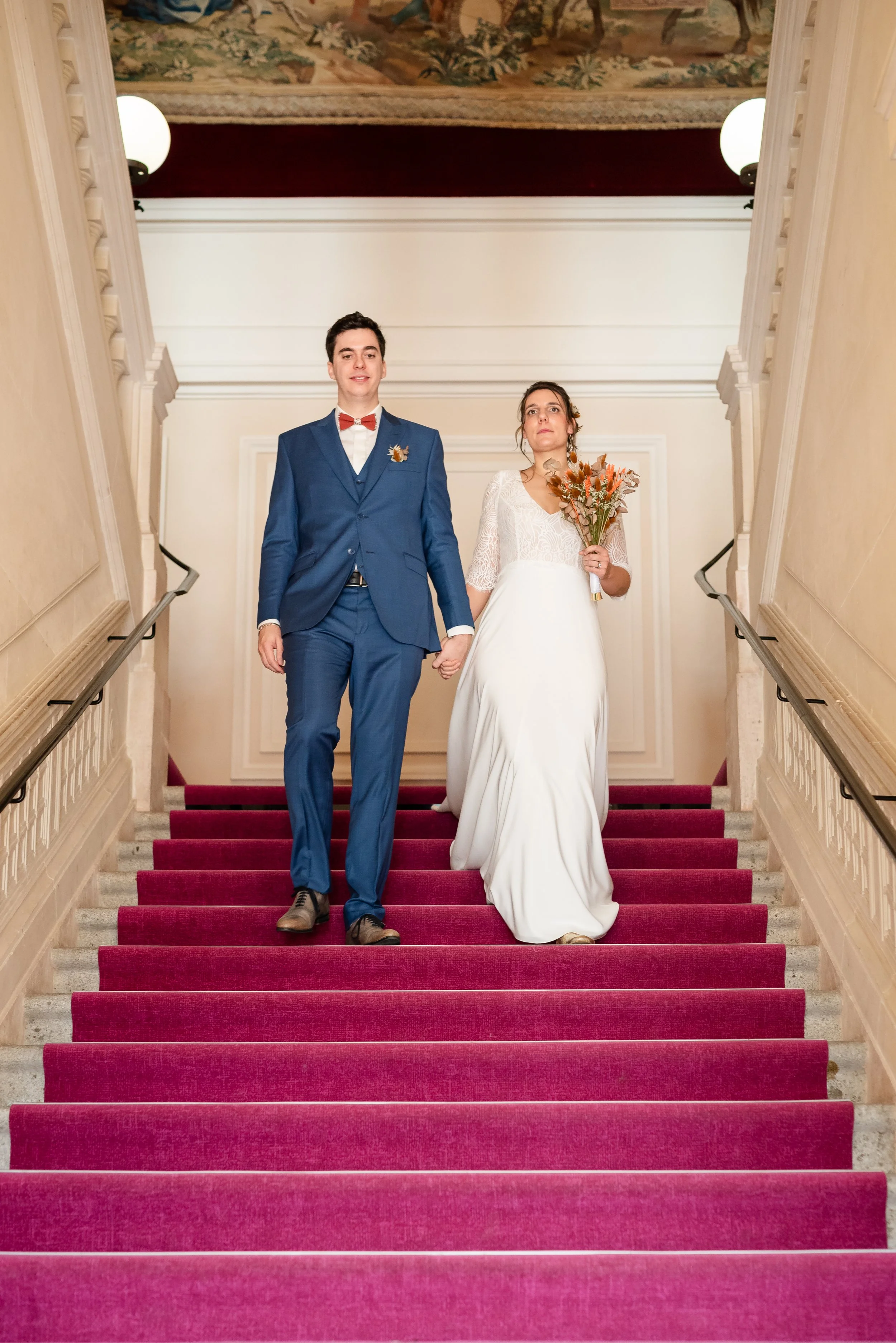 Un couple en mariage, marchant main dans la main sur un escalier avec tapis rouge dans une salle élégante. L'homme porte un costume bleu avec une chemise blanche et un nœud papillon rouge, la femme porte une robe de mariée blanche et tient un bouquet
