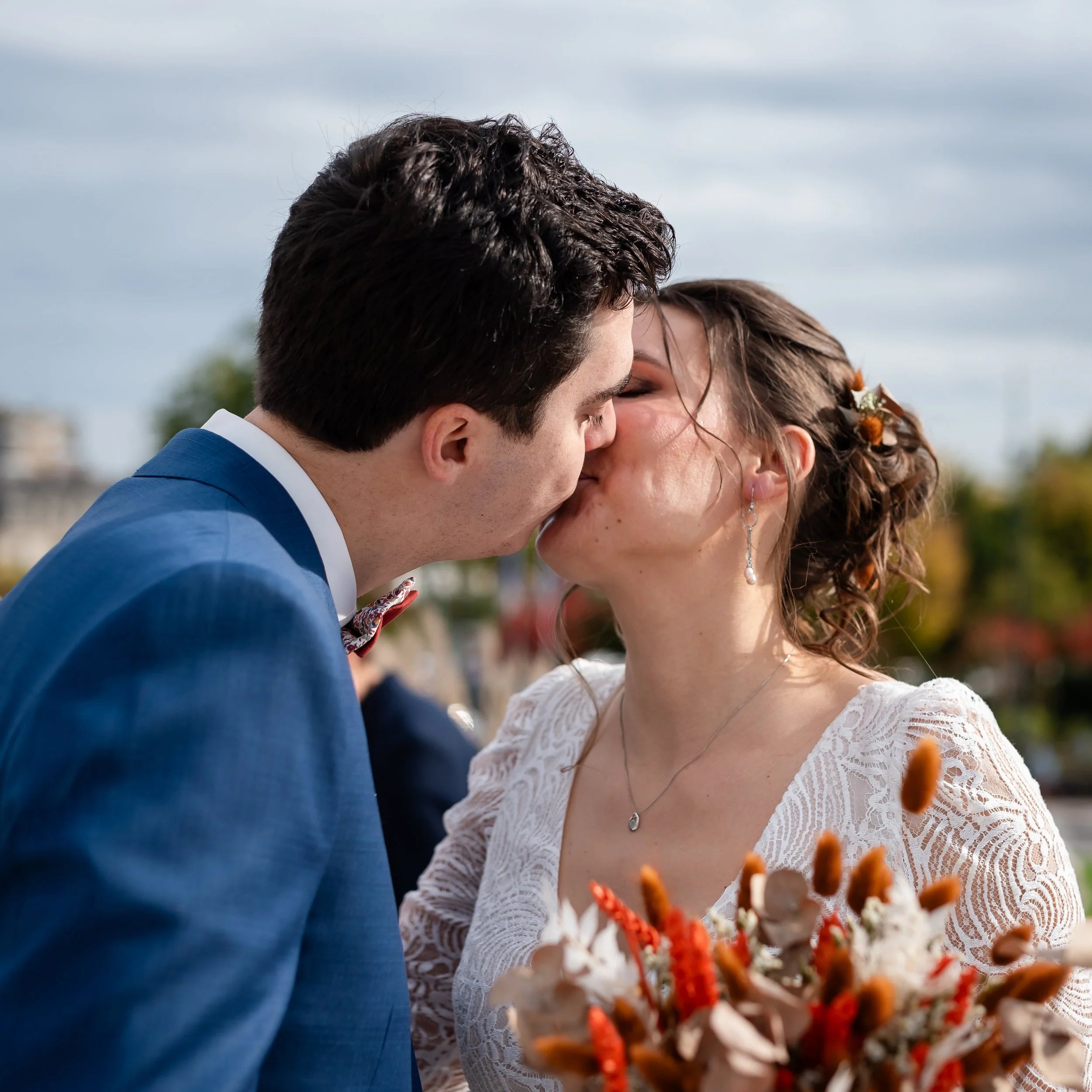 Un couple embrassé lors d'une cérémonie de mariage en extérieur, avec des fleurs dans l'avant-plan et un ciel nuageux en arrière-plan.