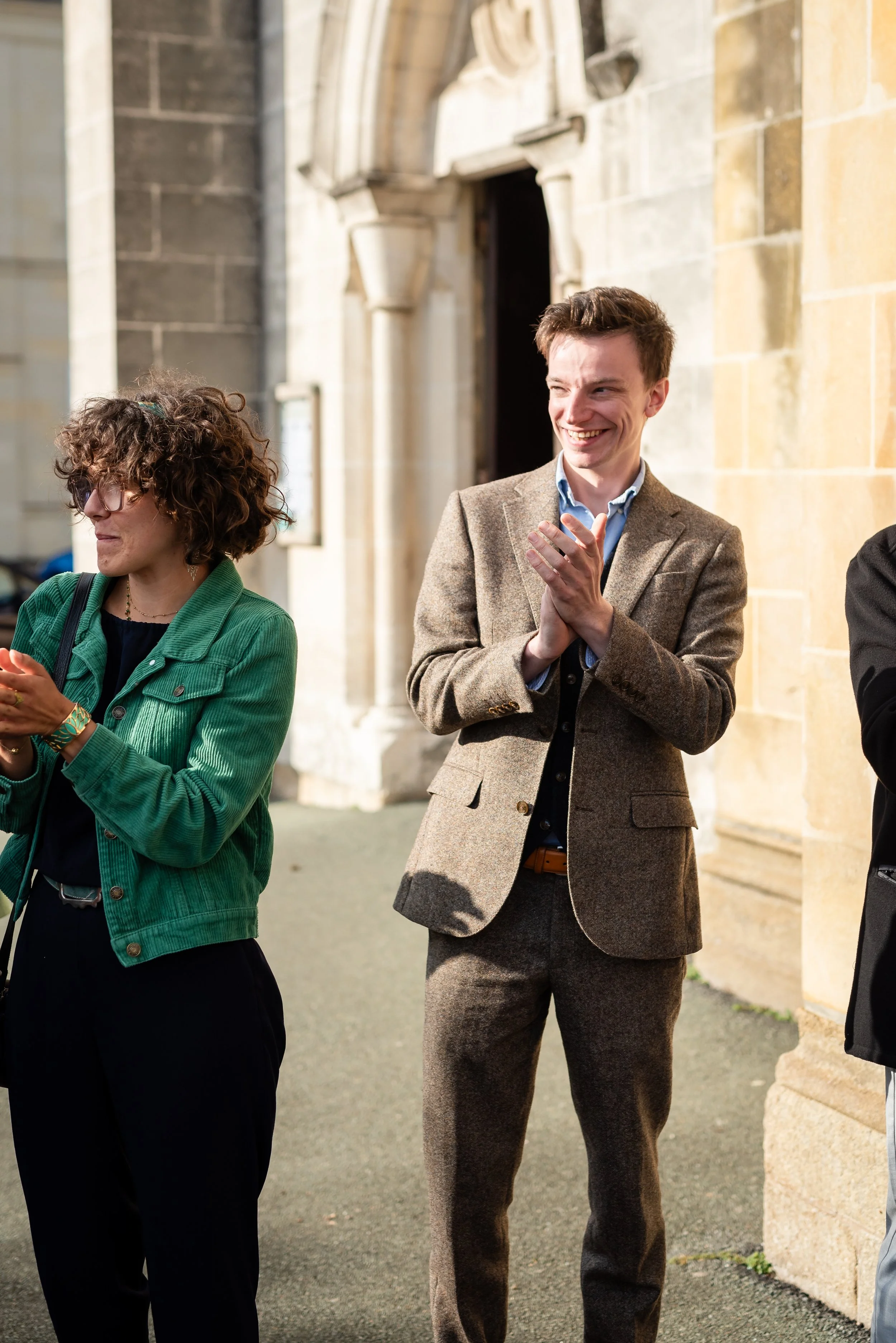 Deux personnes habillées formellement, un homme souriant avec un costume marron et une femme portant une veste verte, debout devant un bâtiment en pierre.