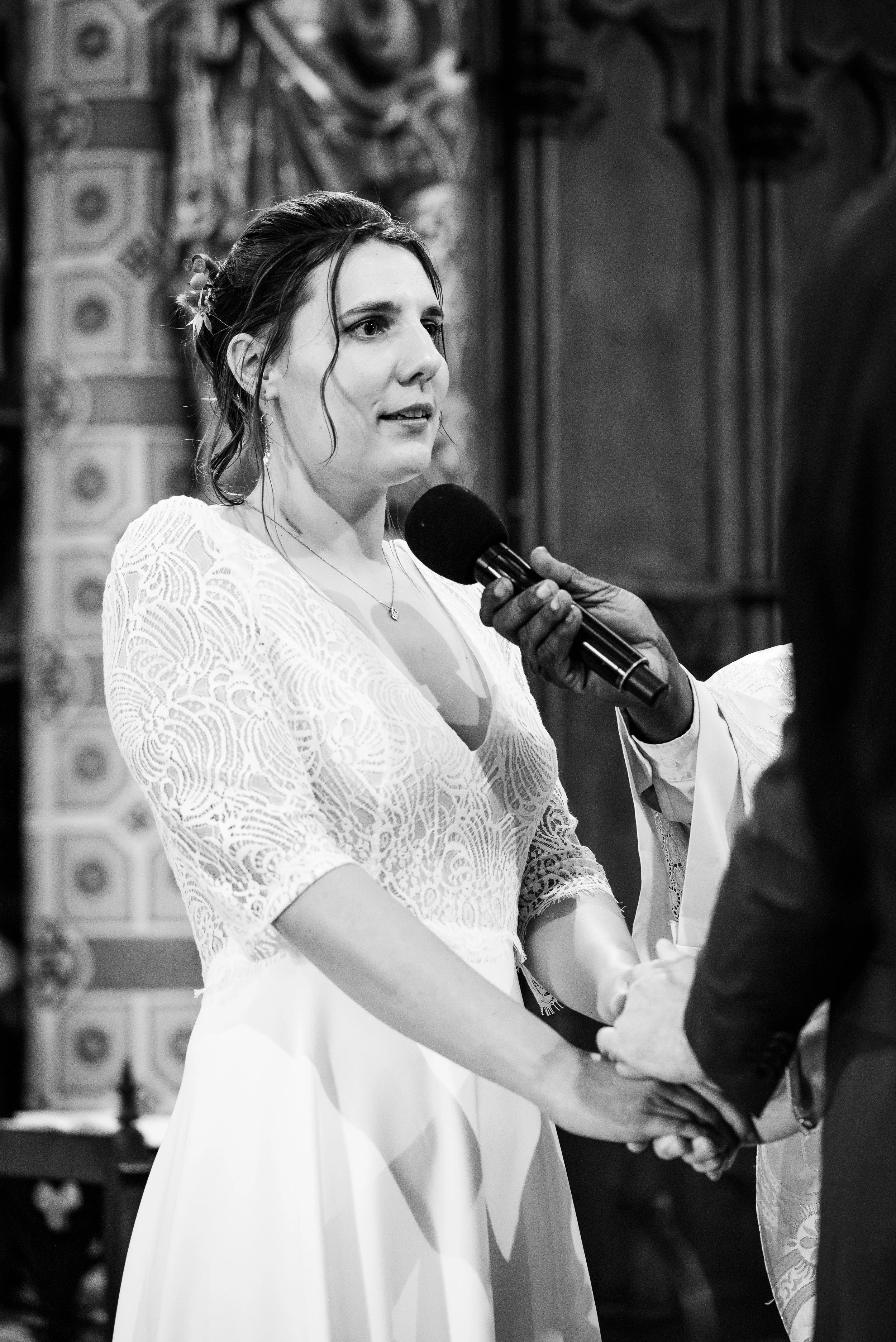 Une femme lors d'une cérémonie, vêtue d'une robe blanche en dentelle et recevant une bénédiction, tenant la main d'une autre personne, dans une ambiance religieuse.