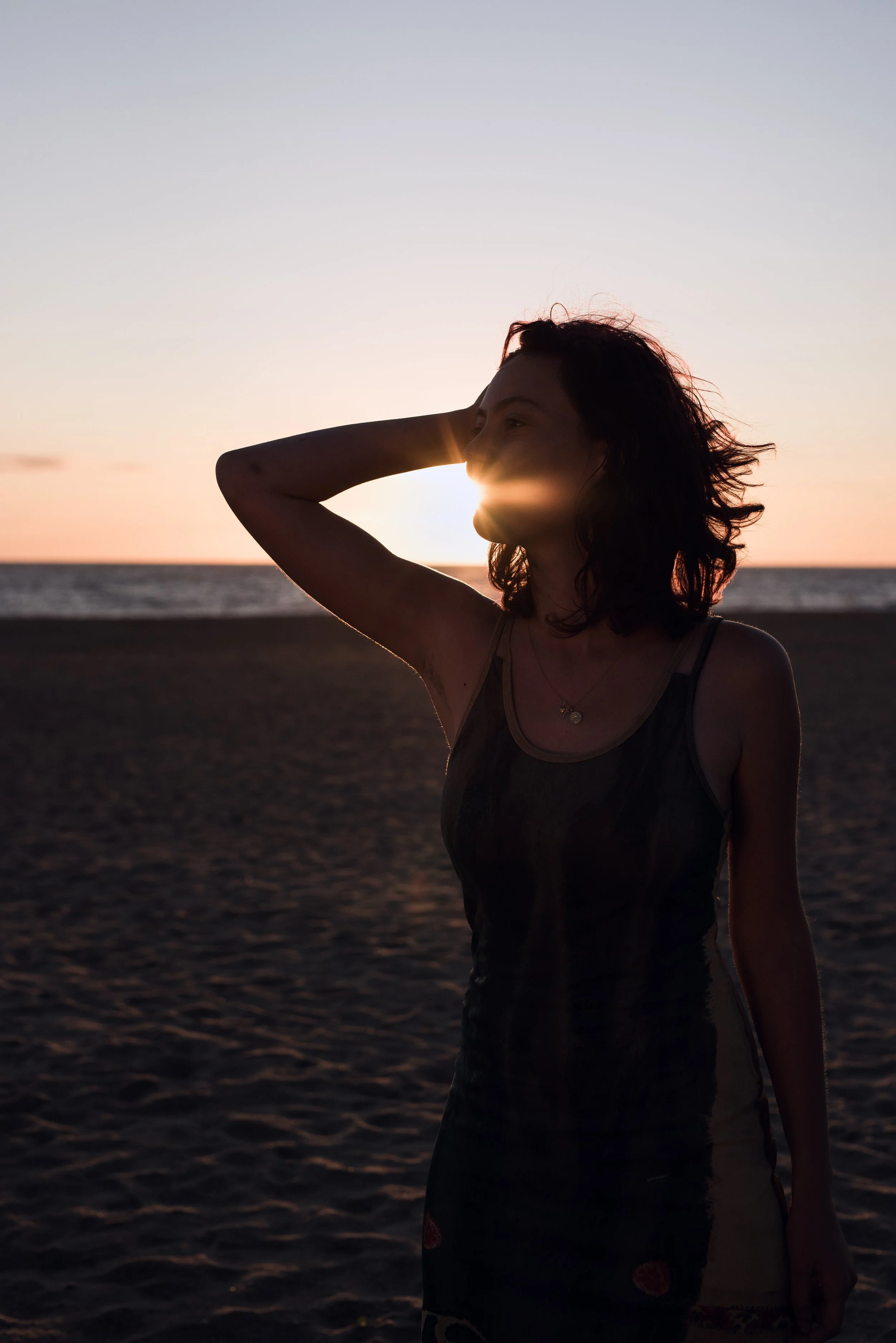 Femme debout sur la plage au coucher du soleil, avec le soleil invisible derrière son visage, regardant vers l'horizon, cheveux bouclés, portant un débardeur noir avec des motifs.