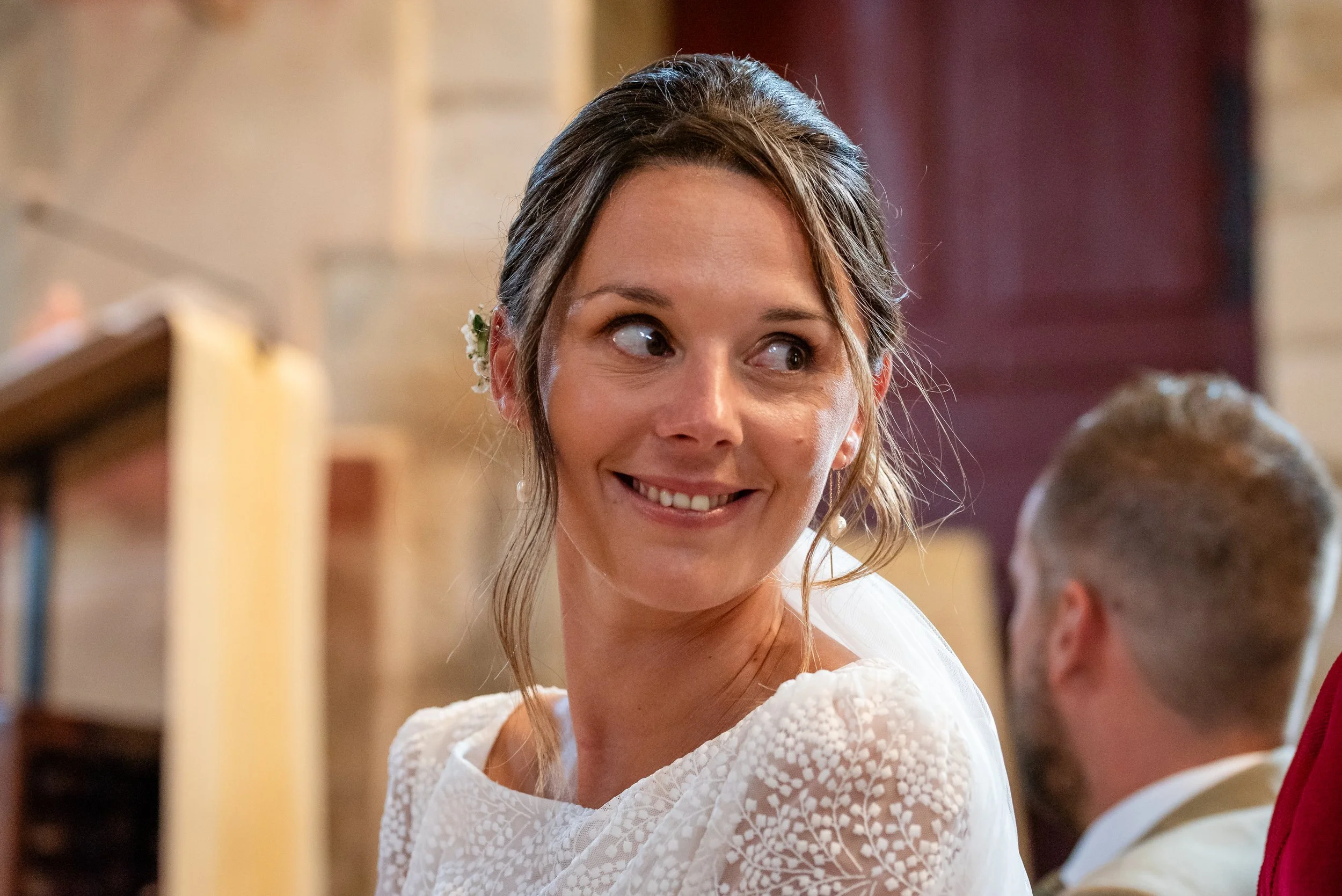 Une femme souriante avec des cheveux attachés, portant une robe blanche en dentelle, à l'intérieur d'un lieu éclairé et décoré, en interaction avec un enfant dont la tête est partiellement visible.