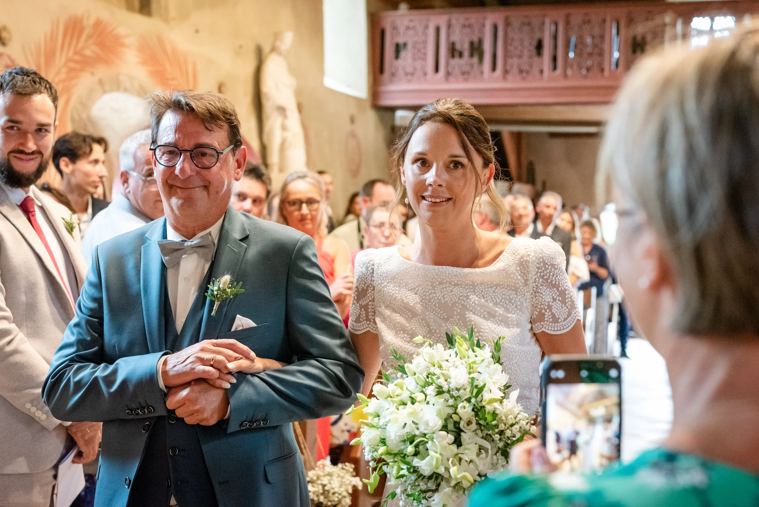 Un couple lors d'un mariage, avec l'homme portant un costume bleu et la femme une robe blanche, entourés d'invités dans une salle de mariage.