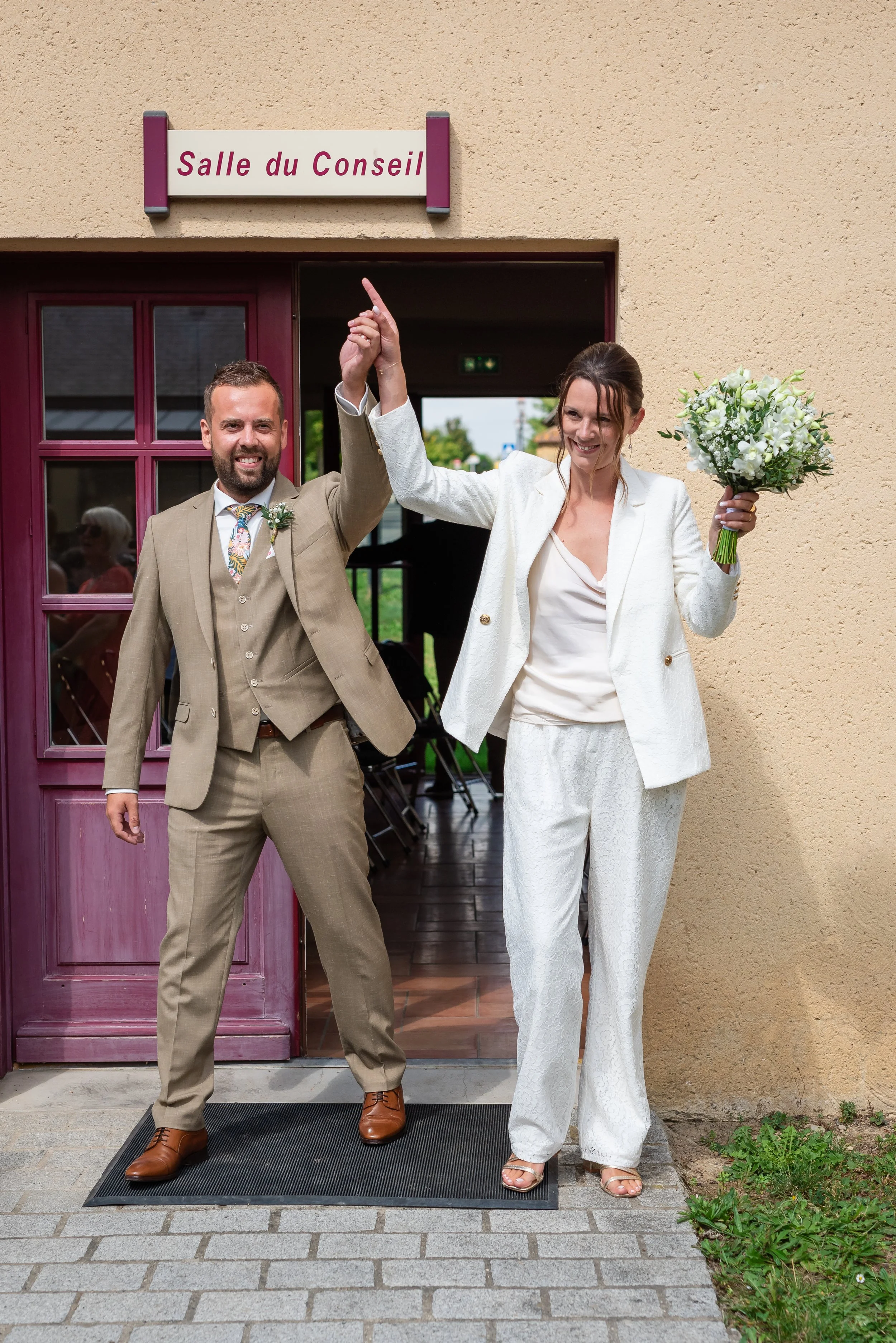 Un homme et une femme en tenue de mariage sortent d'une salle de conseil, se tenant par la main levée, devant une porte violet foncé et un mur beige, la femme tient un bouquet blanc, ils riant et souriant.