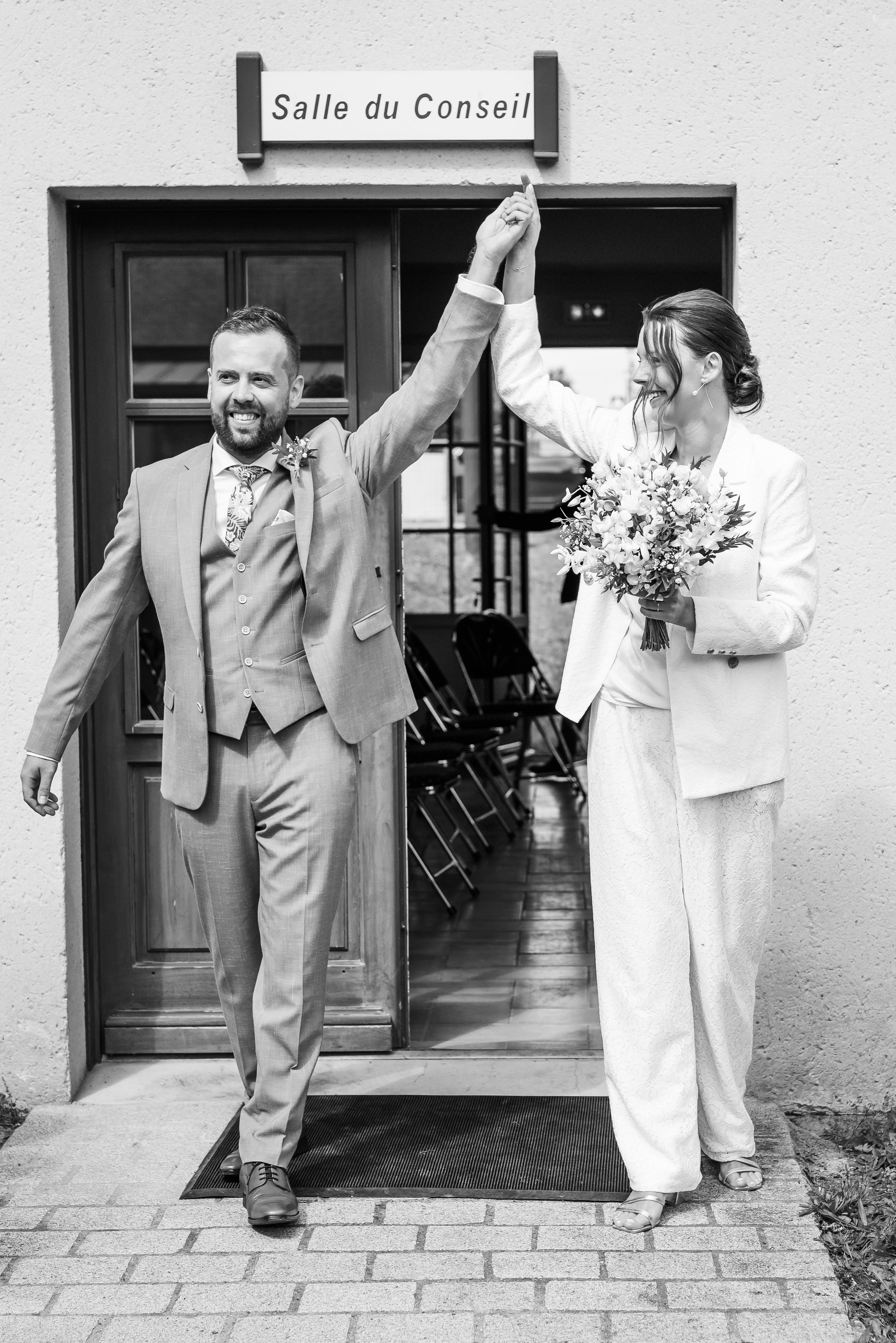 Un couple marié sortant d'une salle de réunion (présumée être la salle du conseil) après leur mariage, se tenant la main en l'air et souriant. La femme tient un bouquet de fleurs dans une main.