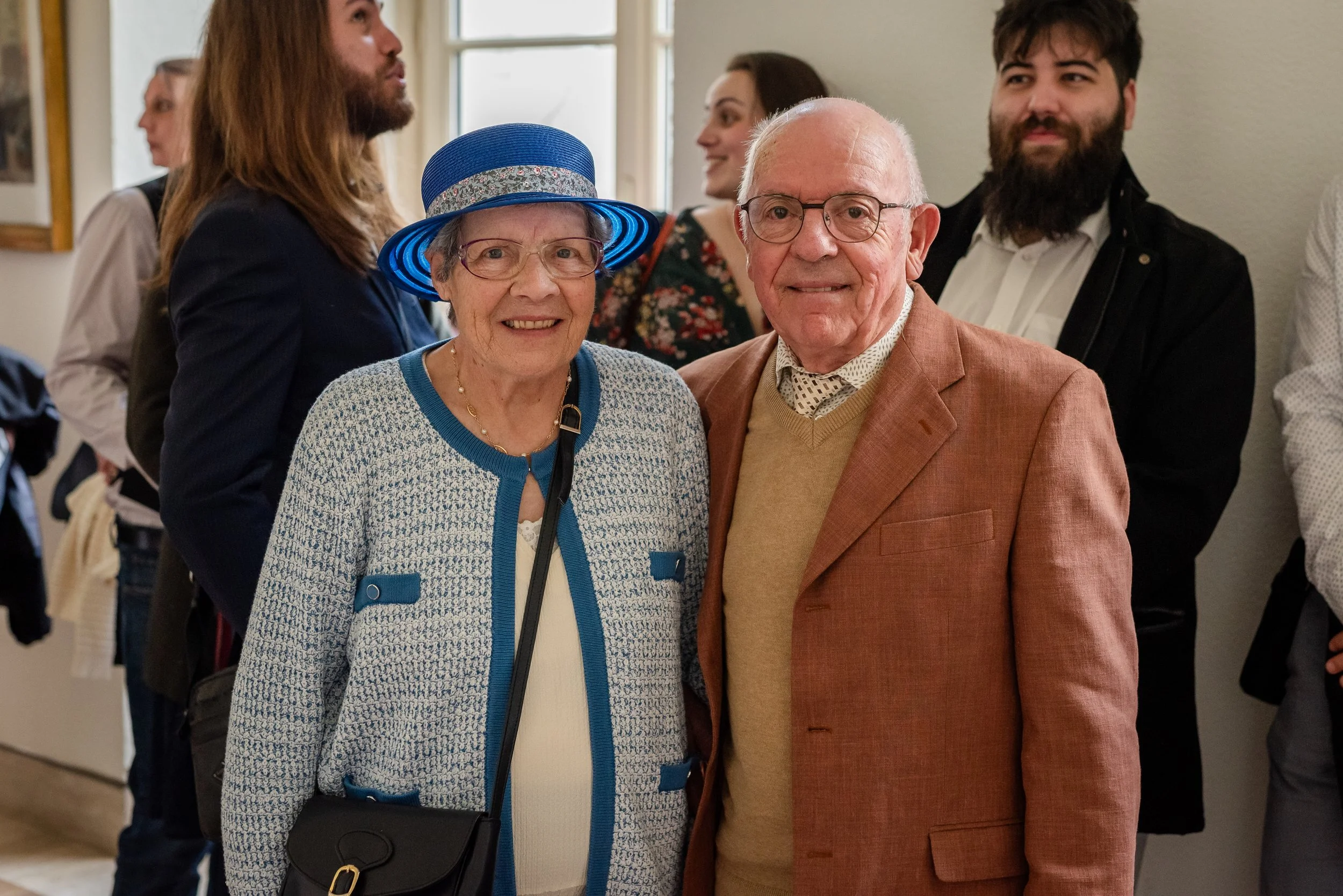 Deux personnes âgées, une femme avec un chapeau bleu et un homme avec des lunettes, posent pour une photo lors d'un événement social, avec des jeunes adultes en arrière-plan.