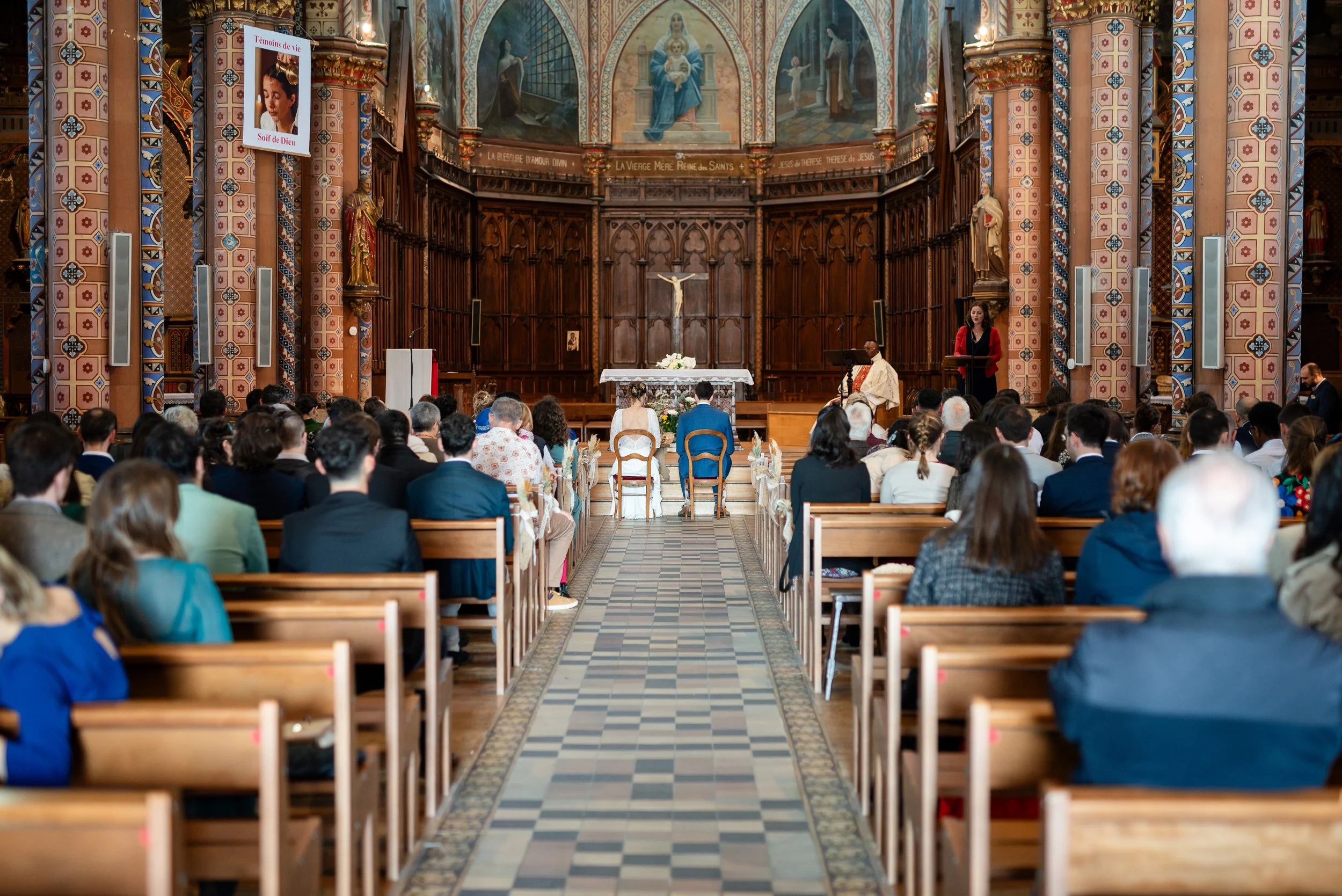 Une cérémonie religieuse dans une église avec des personnes assises, un prêtre, et un couple devant l'autel. L'autel est décoré avec une croix et des fleurs, et l'intérieur de l'église est richement décoré avec des fresques et des statues.