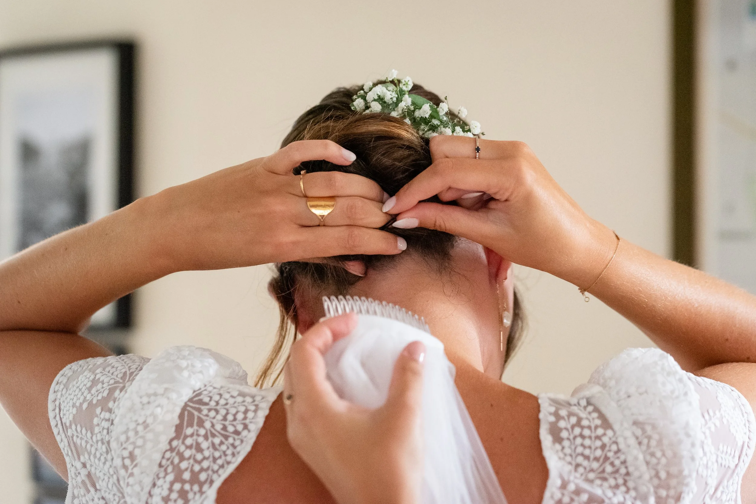 Femme coiffant ses cheveux avec une coiffure en chignon décorée de petites fleurs blanches, portant une robe en dentelle blanche.