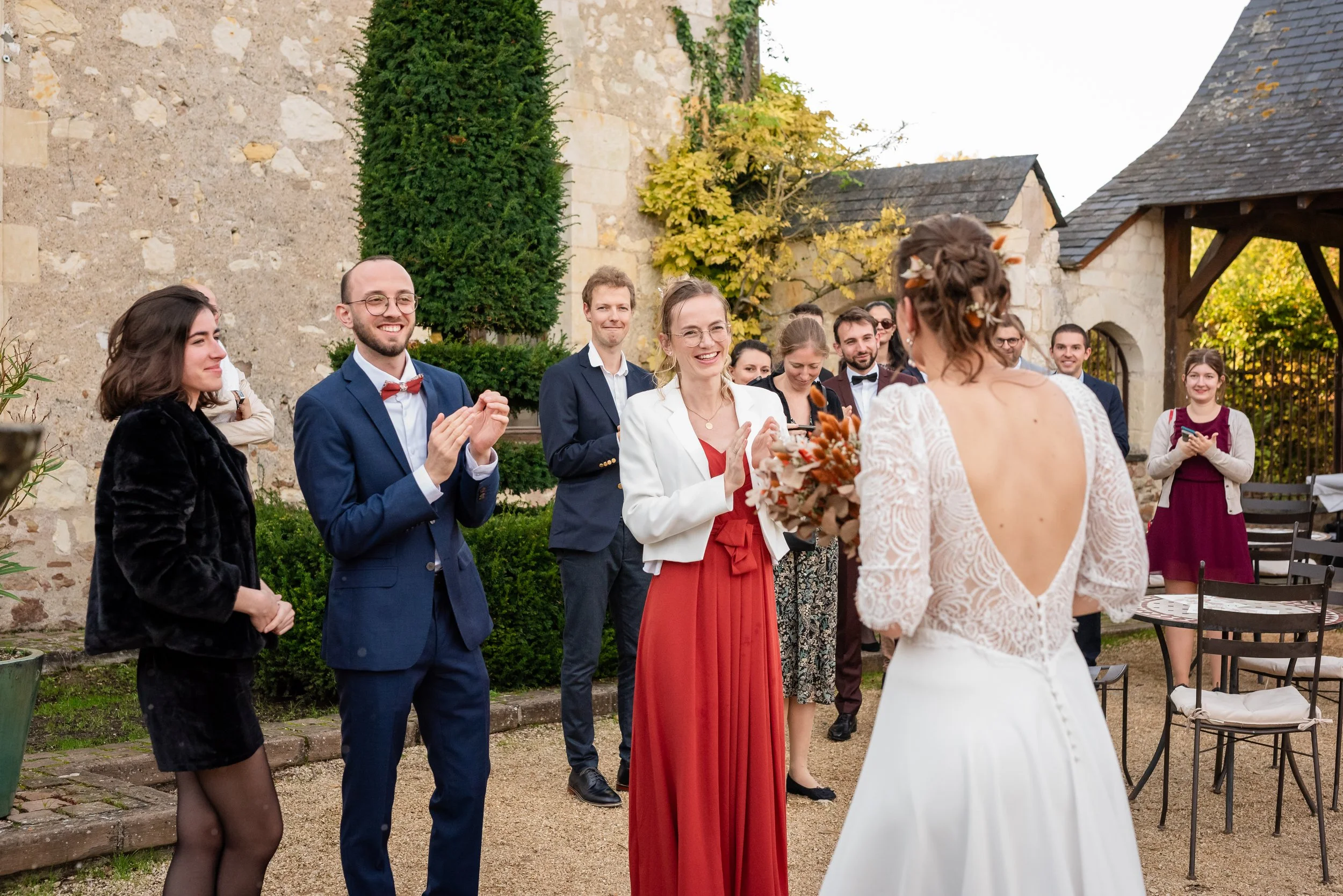 Groupe de personnes lors d'un mariage à l'extérieur, avec la mariée en premier plan et les invités qui l'entourent, dans un cadre romantique avec des murs en pierre et des arbres aux couleurs automnales.